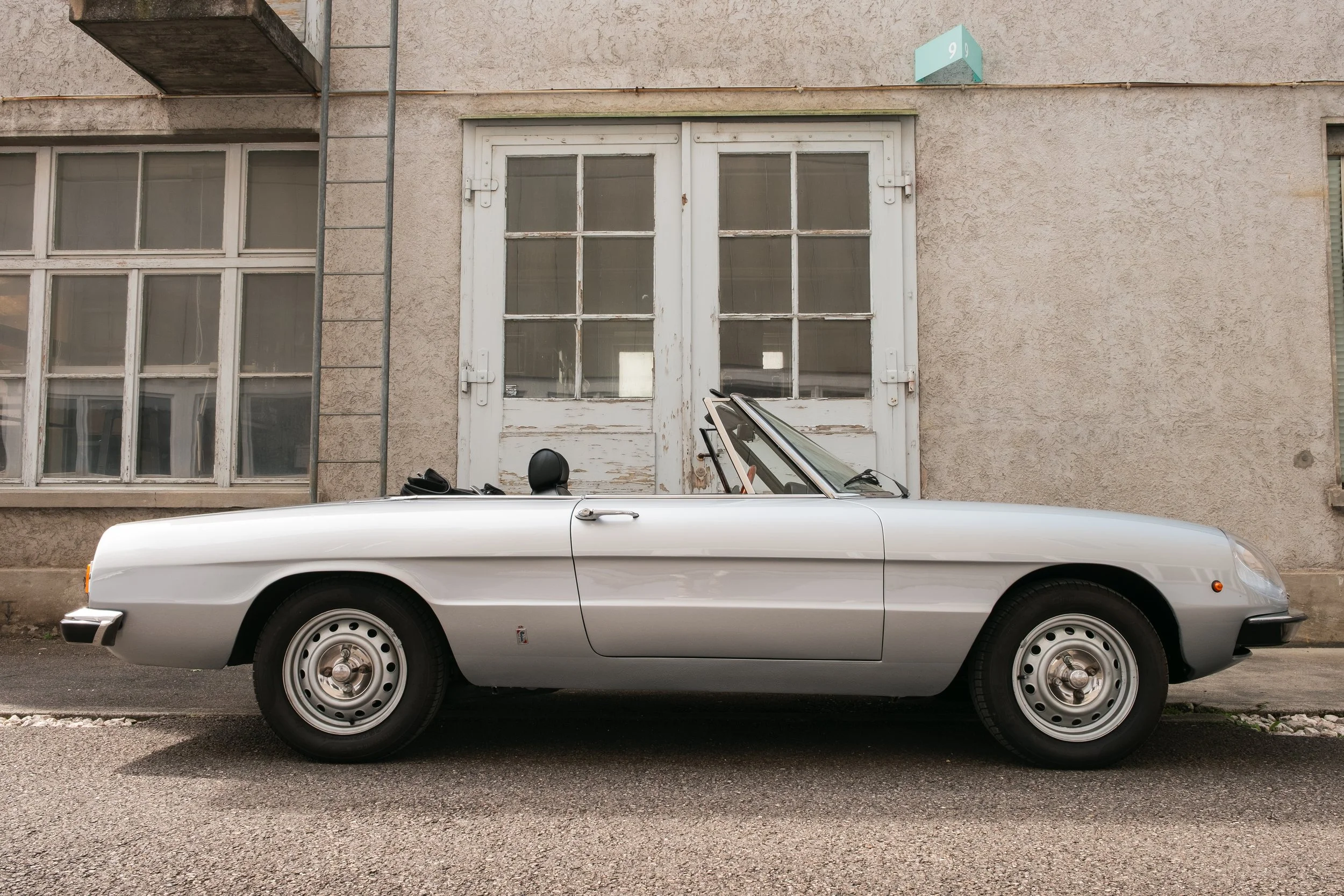 Ein silberner Oldtimer-Cabrio vor einer alten, beige Fassadenwand mit großen, weißen Fenster und doppelflügeligen Türen in einer Stadtstraße.
