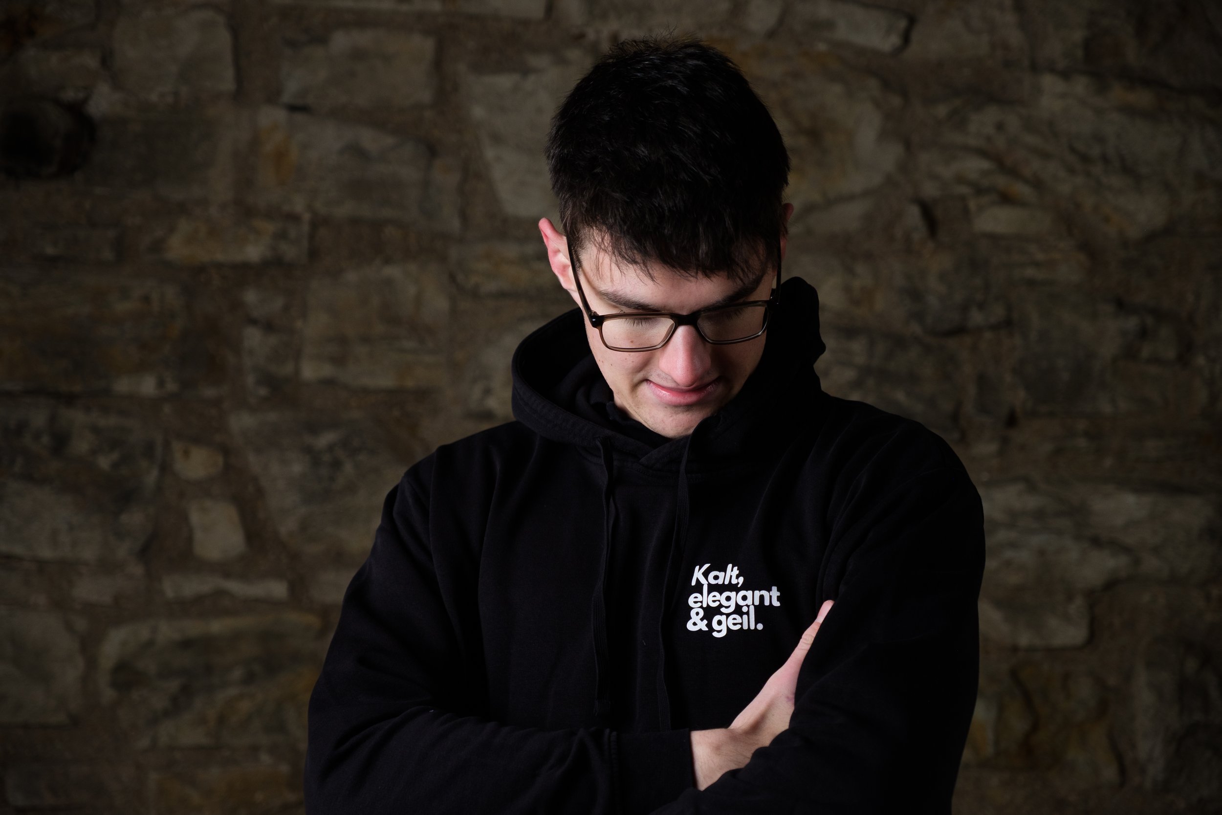 Ein junger Mann mit dunklem Haar und Brille trägt einen schwarzen Hoodie mit dem weißen Aufdruck „Kalt, elegant & geil.“ und steht vor einer steinernen Wand, während er nach unten blickt.