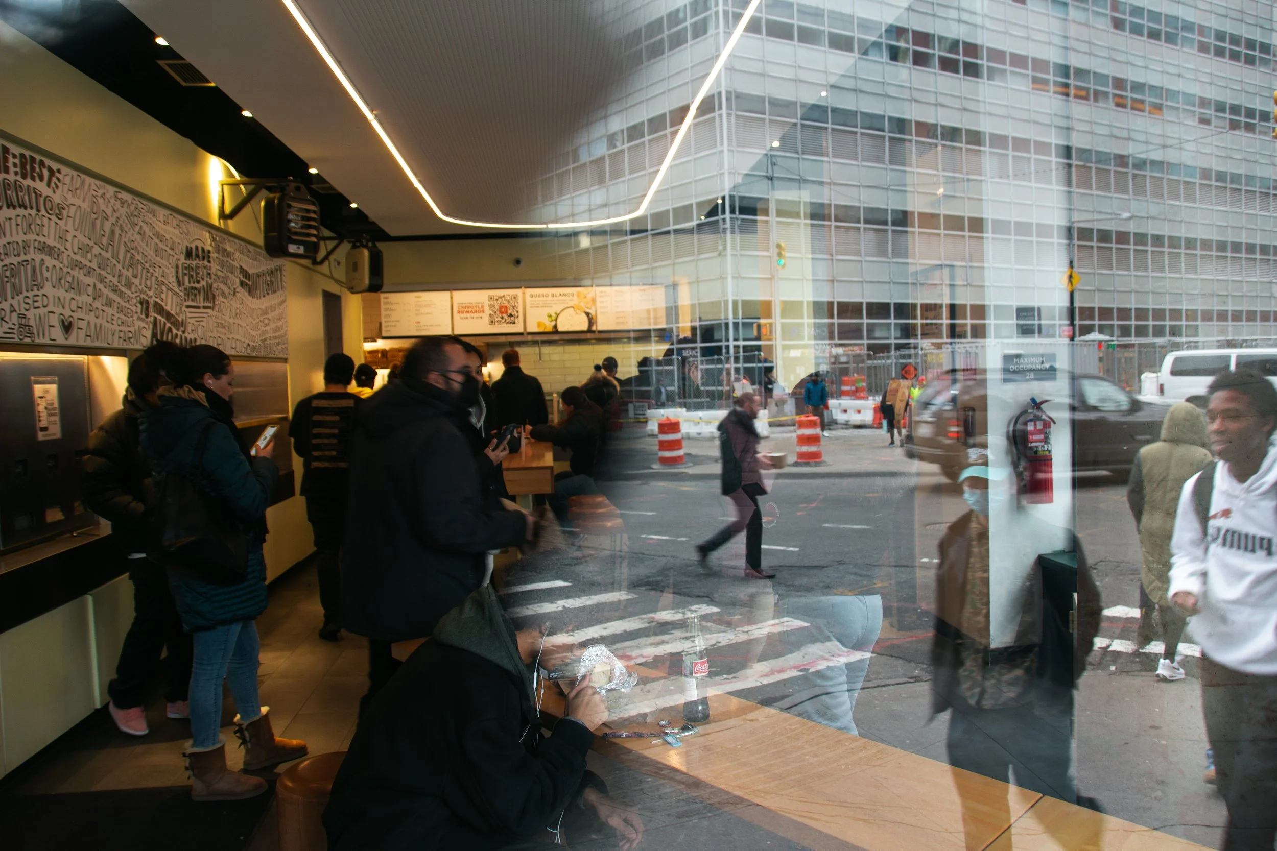 Menschen im Café und draußen auf der Straße in einer Zeitrafferaufnahme, Blick durch eine Glasfront auf den Straßenverkehr und Fußgänger, reflektierende Fassaden im Hintergrund.