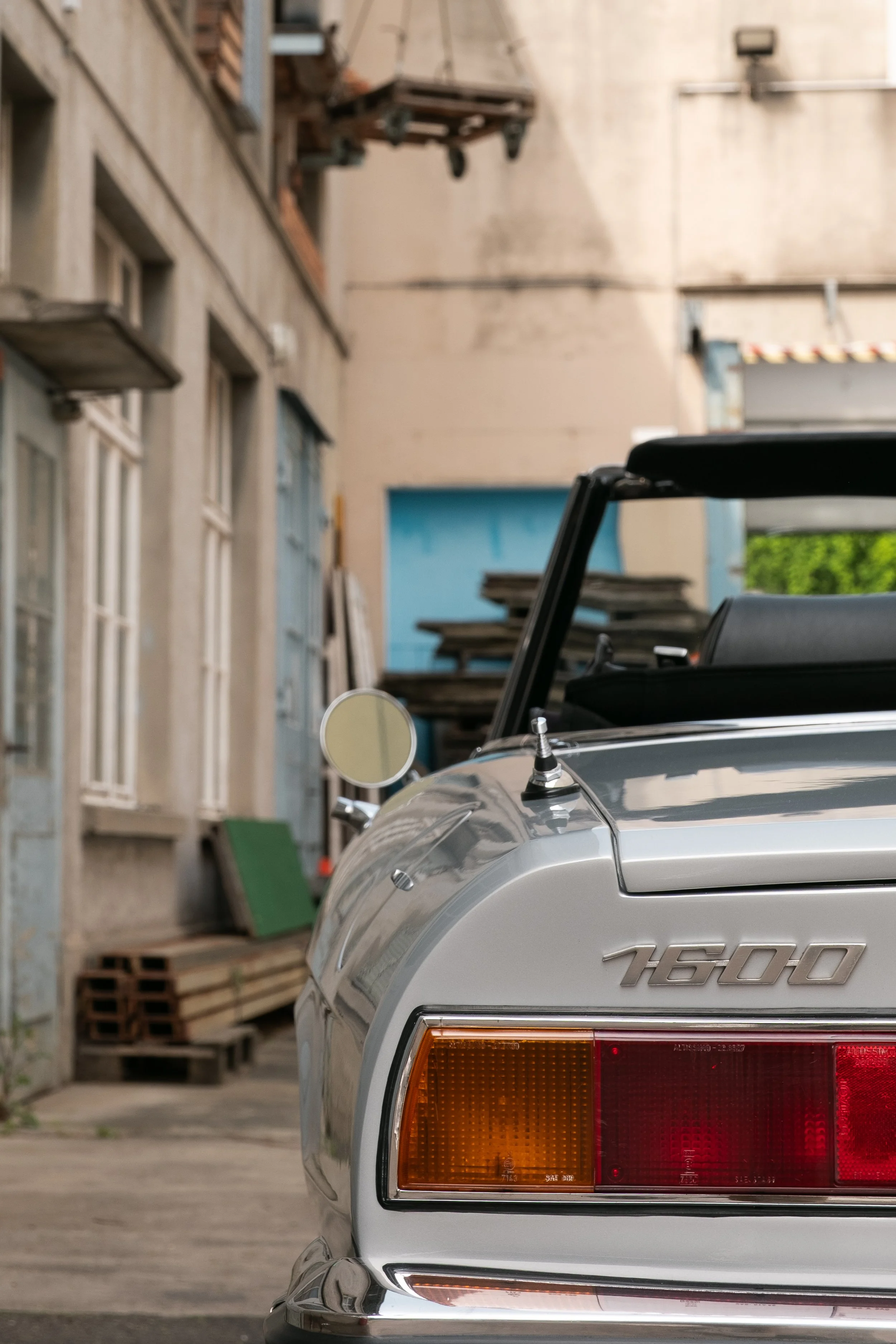 Ein silberner Sportwagen, Modell 1600, steht in einer Garage oder Werkstatt. Im Hintergrund sind abgenutzte Gebäude, Holzstücke und eine gelbe Wand sichtbar.
