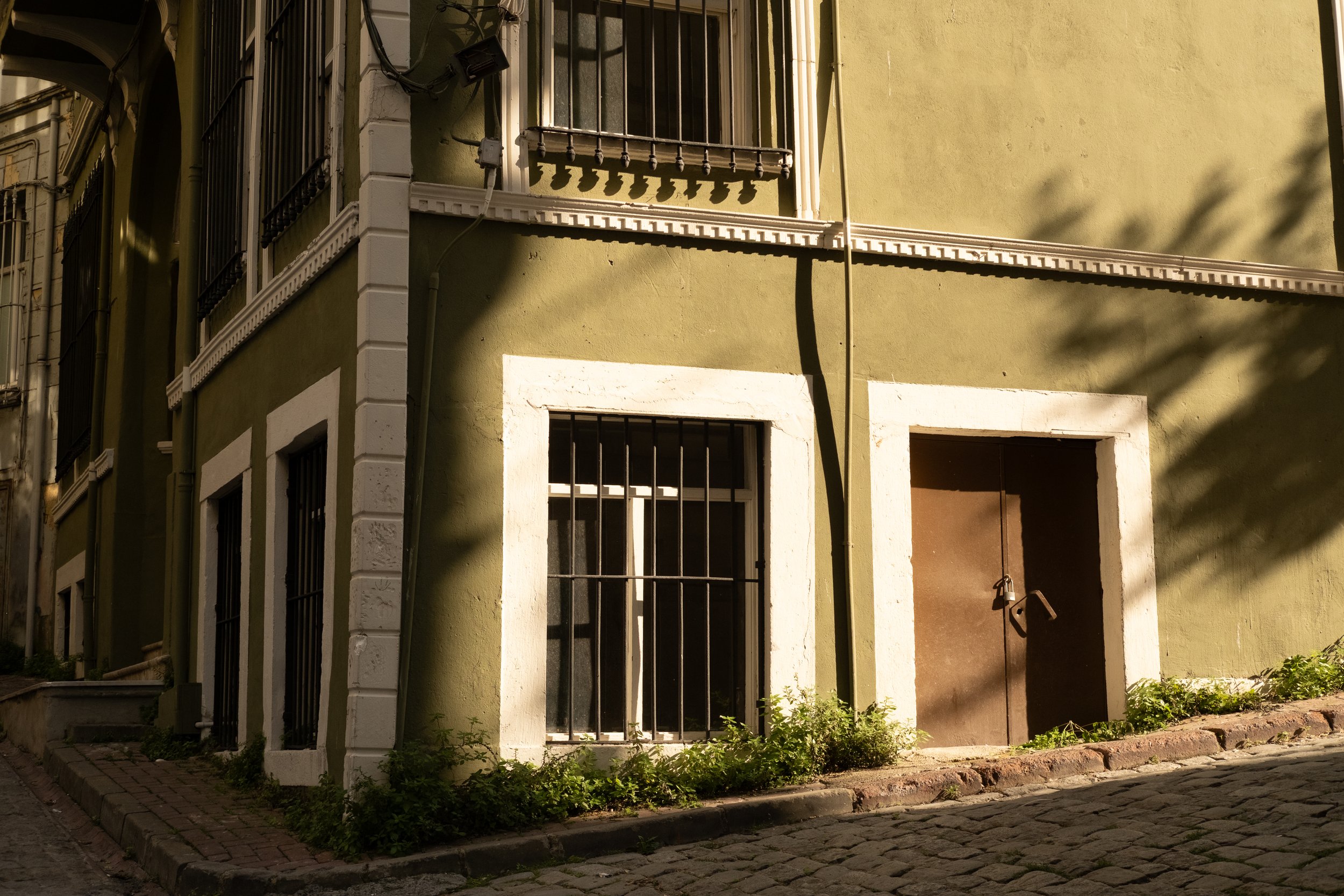 Ecke eines grünen Gebäudes mit weißen Rahmen um Fenster und Tür, mit Gittergeschäften auf den Fenstern, an einer Kopfsteinpflasterstraße, mit Schatten von Bäumen