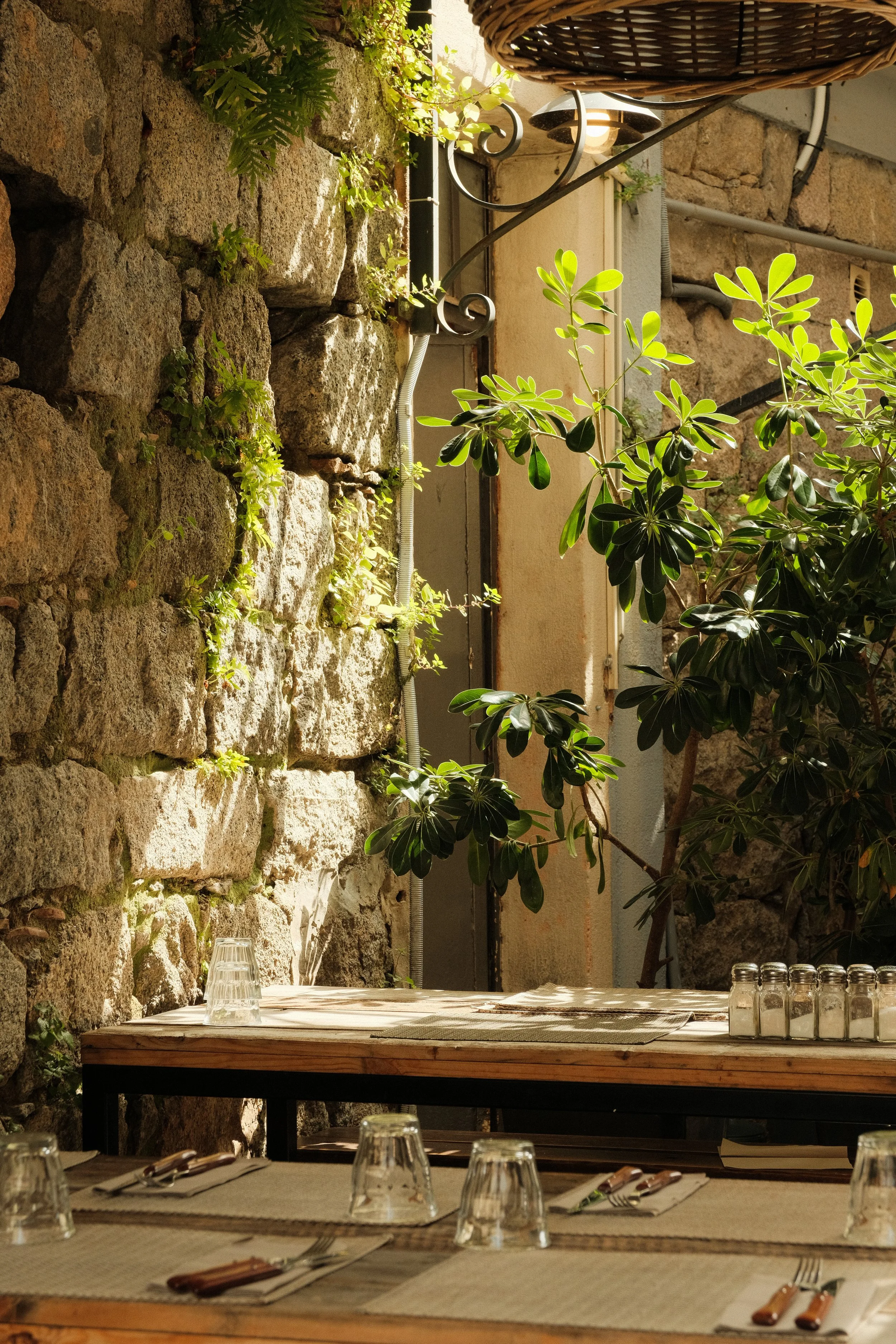 Ein gemütlicher Außenbereich eines Restaurants oder Cafés mit einem Holztisch, Besteck, Gläsern, einer Steinmauer, grünen Pflanzen und Schatten spenden.