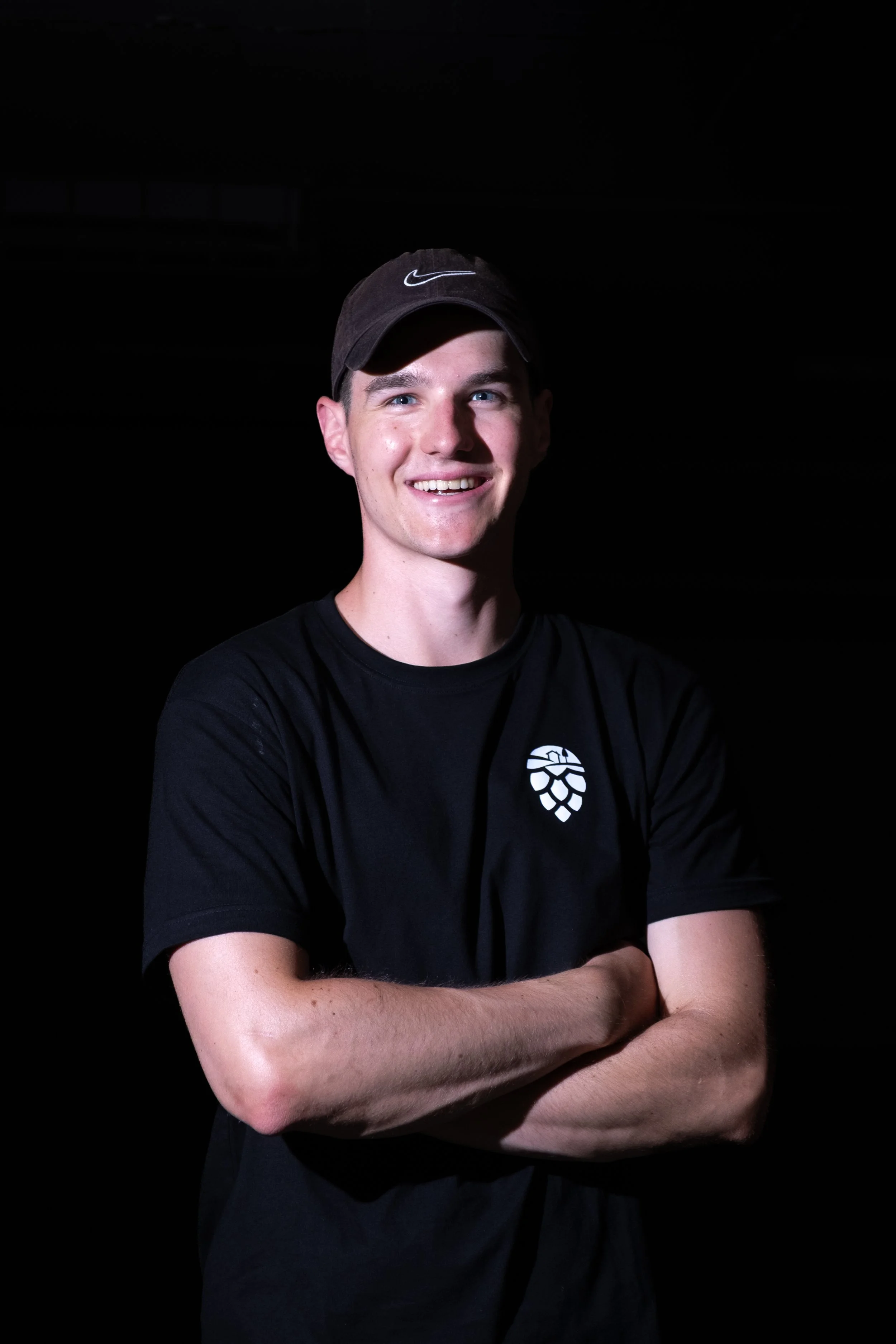 Junger Mann mit schwarzem T-Shirt und Baseballcap, lächelnd und die Arme verschränkt, vor schwarzem Hintergrund.