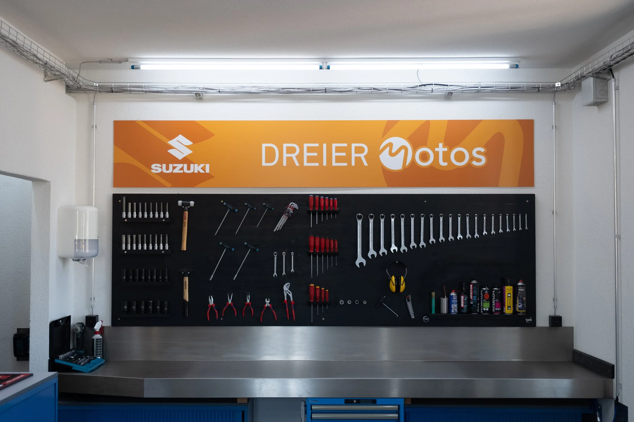Werkzeugwand mit verschiedenen Schraubenschlüsseln, Schraubenziehern, Zangen und Werkstattmaterialien in einer Autowerkstatt, mit einem orangefarbenen Werbeträger von Suzuki und Dreier Motos an der Wand.