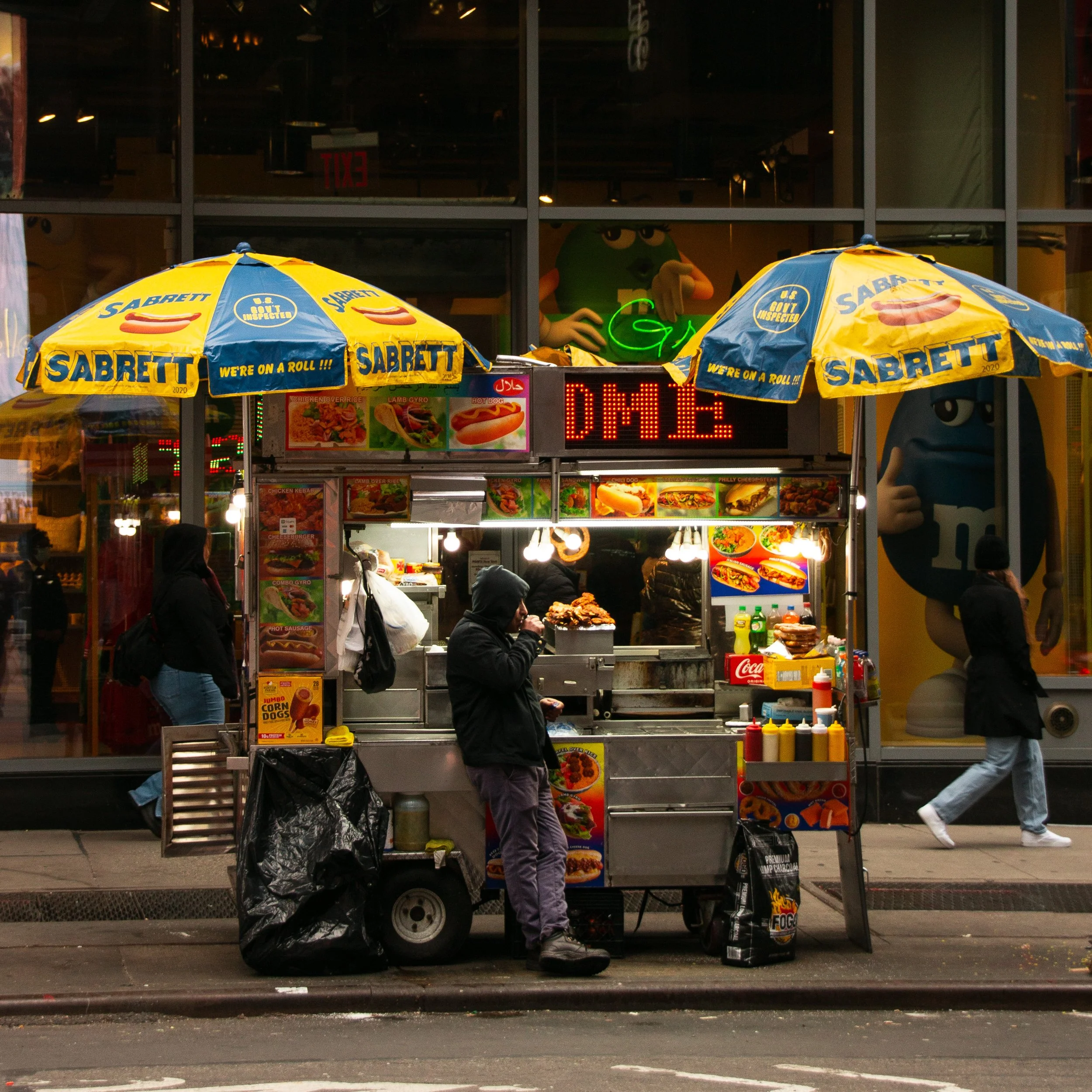 Straßenimbissstand mit Cook-Wagen, Essensangeboten und bunten Schirmen, Menschen in dunkler Kleidung, Neonbeschilderung und M&M's-Werbung im Hintergrund.