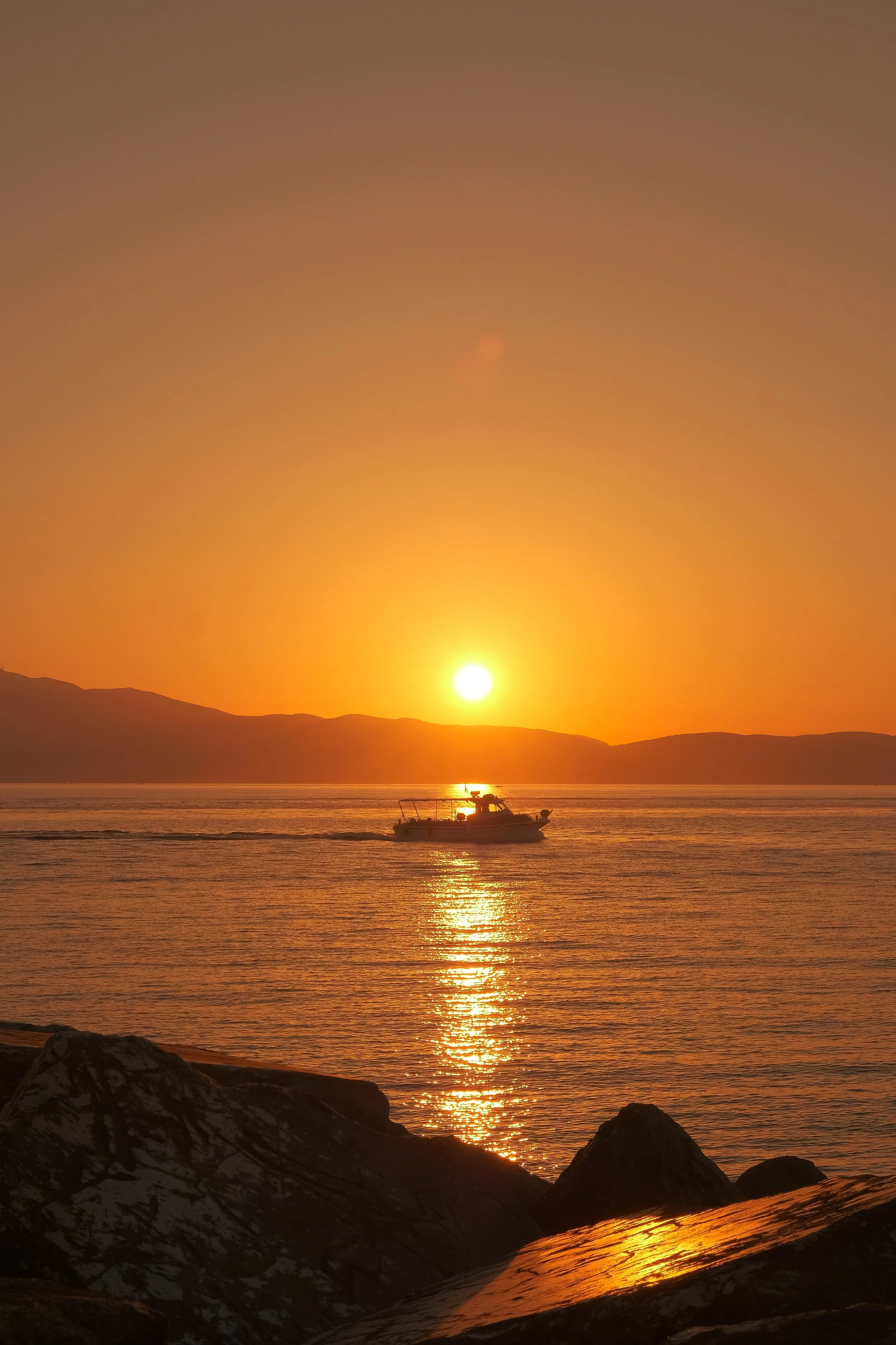 Ein Sonnenuntergang über einem Wasser, mit einem Boot auf dem Wasser und Felsen im Vordergrund.