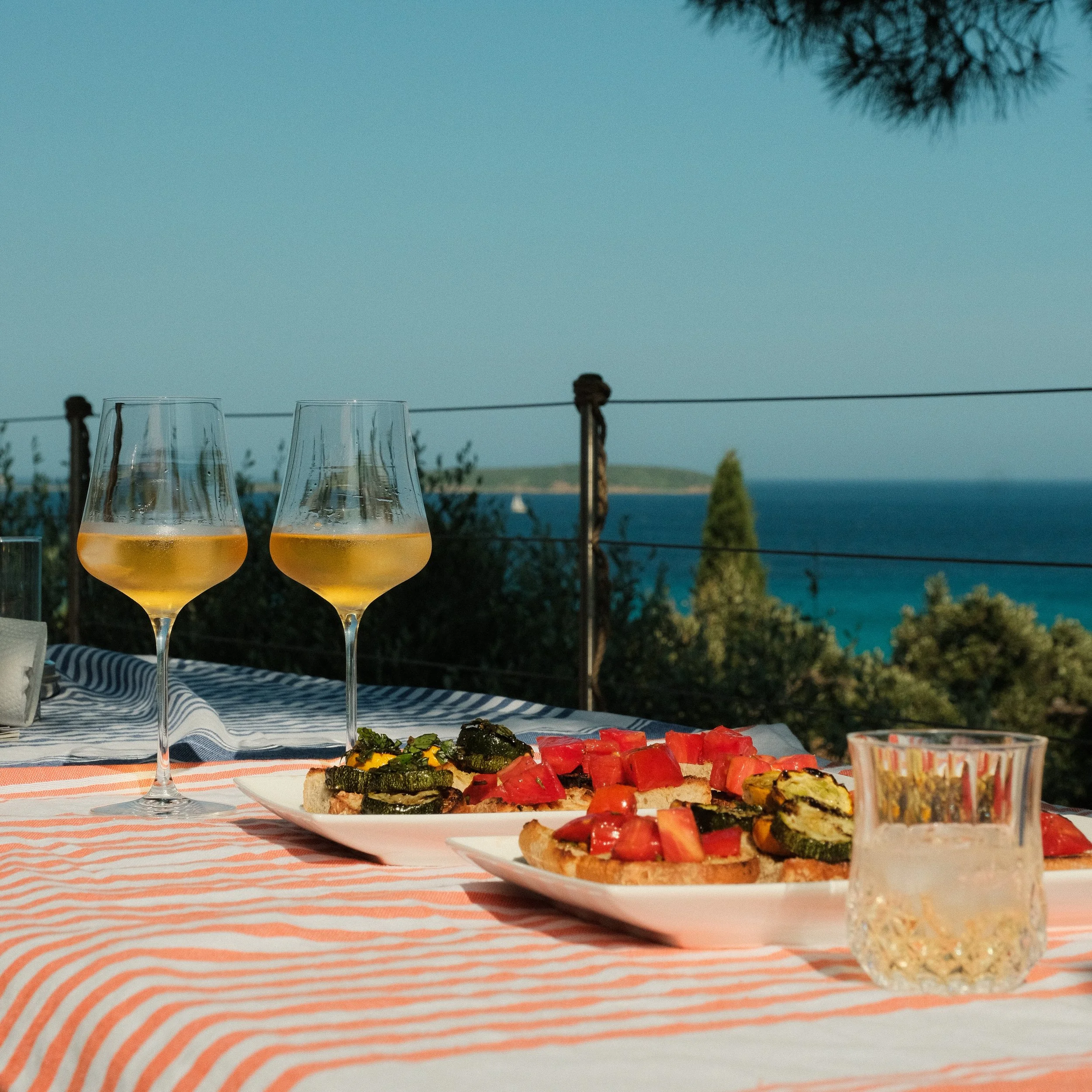 Ein Tisch mit zwei Gläsern weißwein, zwei kleinen Platten mit gegrilltem Gemüse und Tomaten, ein Glas Wasser, im Hintergrund Blick auf das Meer und einen blauen Himmel.