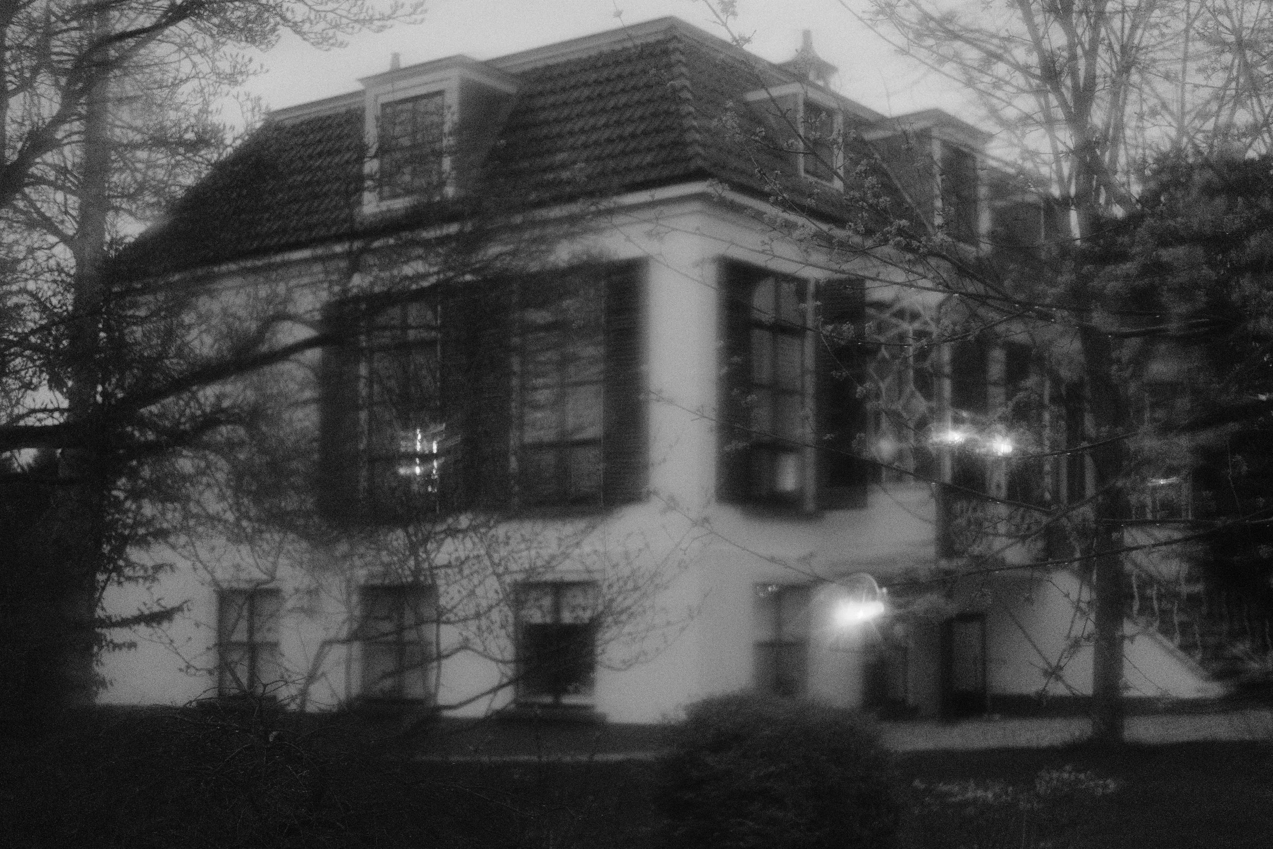 Ein mehrstöckiges Haus mit dunklem Dach, umgeben von Bäumen, die im Winter blattlos sind, bei Nacht, mit Straßenlaternen, die Licht auf das Haus werfen.