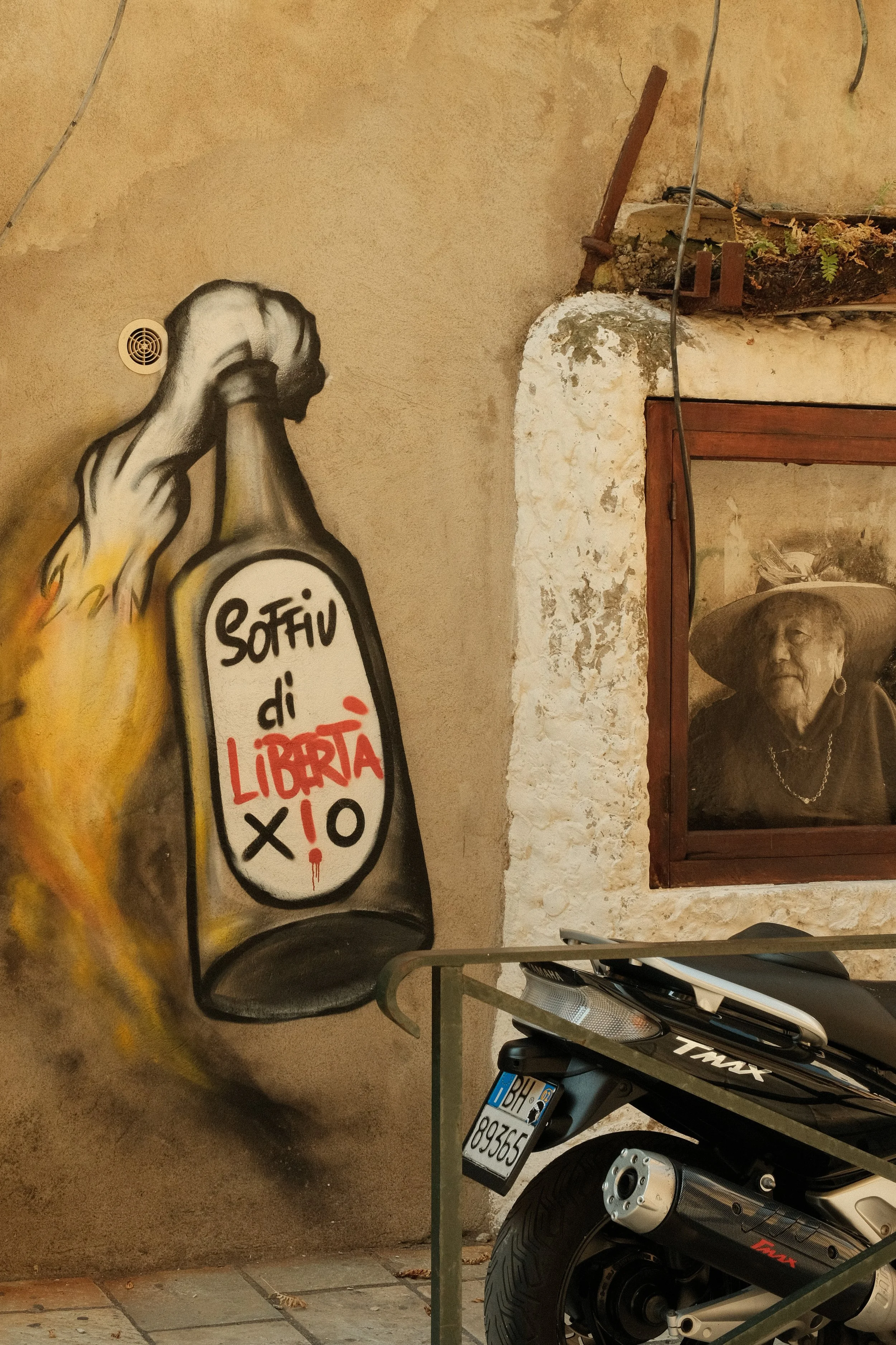 Straßenkunst mit einem Spray-Graffiti eines Hand mit einer Flasche, auf der steht 'Sofiv di libertà io' in Schwarz, Rot und Weiß, an einer alten Wand neben einem Motorrad und einem Fenster mit einem alten Mädchen in einem großen Hut.