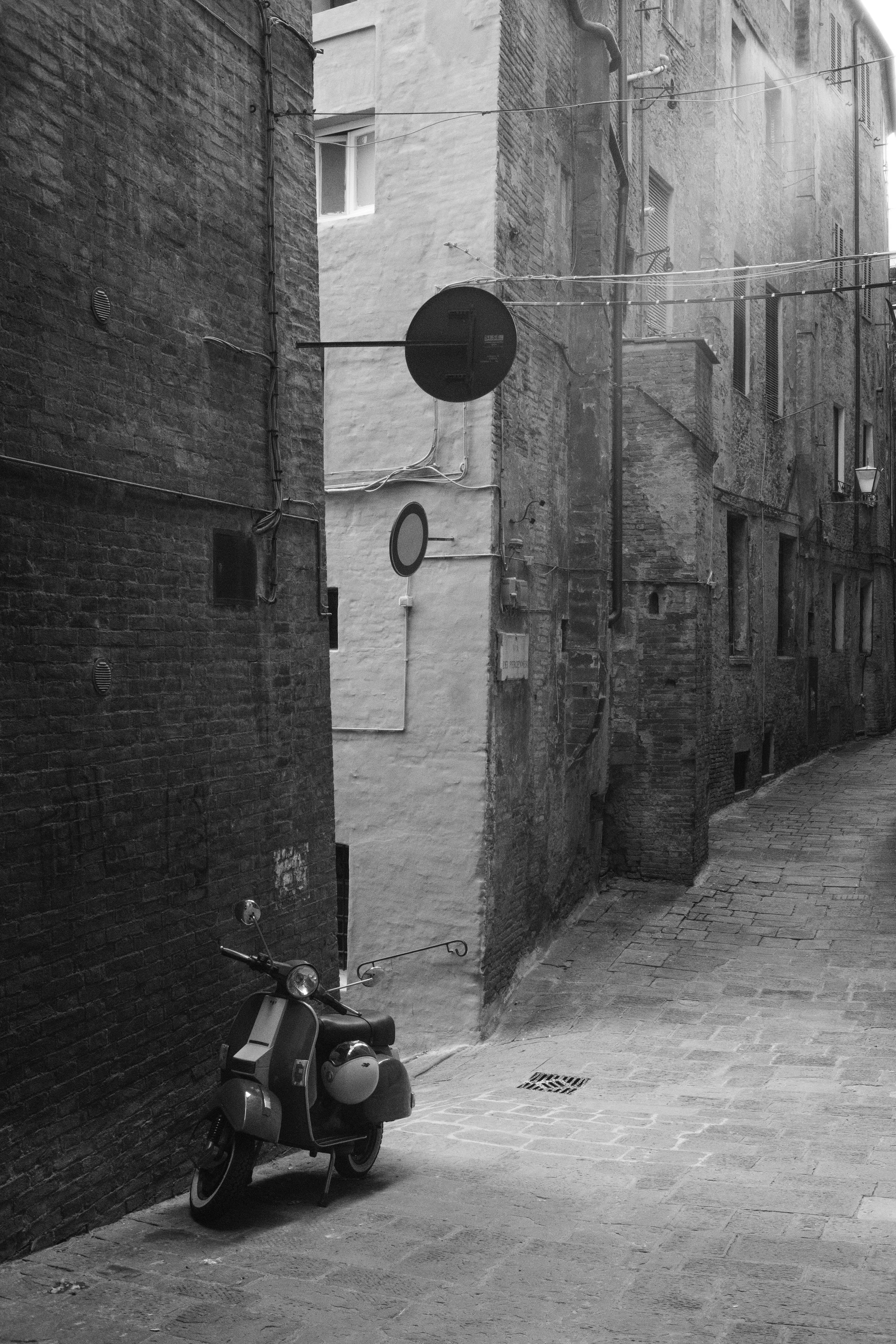 Eine enge, kurvige Gasse mit alten, aneinander gereihten Backstein- und Putzhäusern. Ein rollendes Vespa steht an der Wand, und mehrere Kabel hängen über der Straße. Es ist eine Schwarz-Weiß-Aufnahme.
