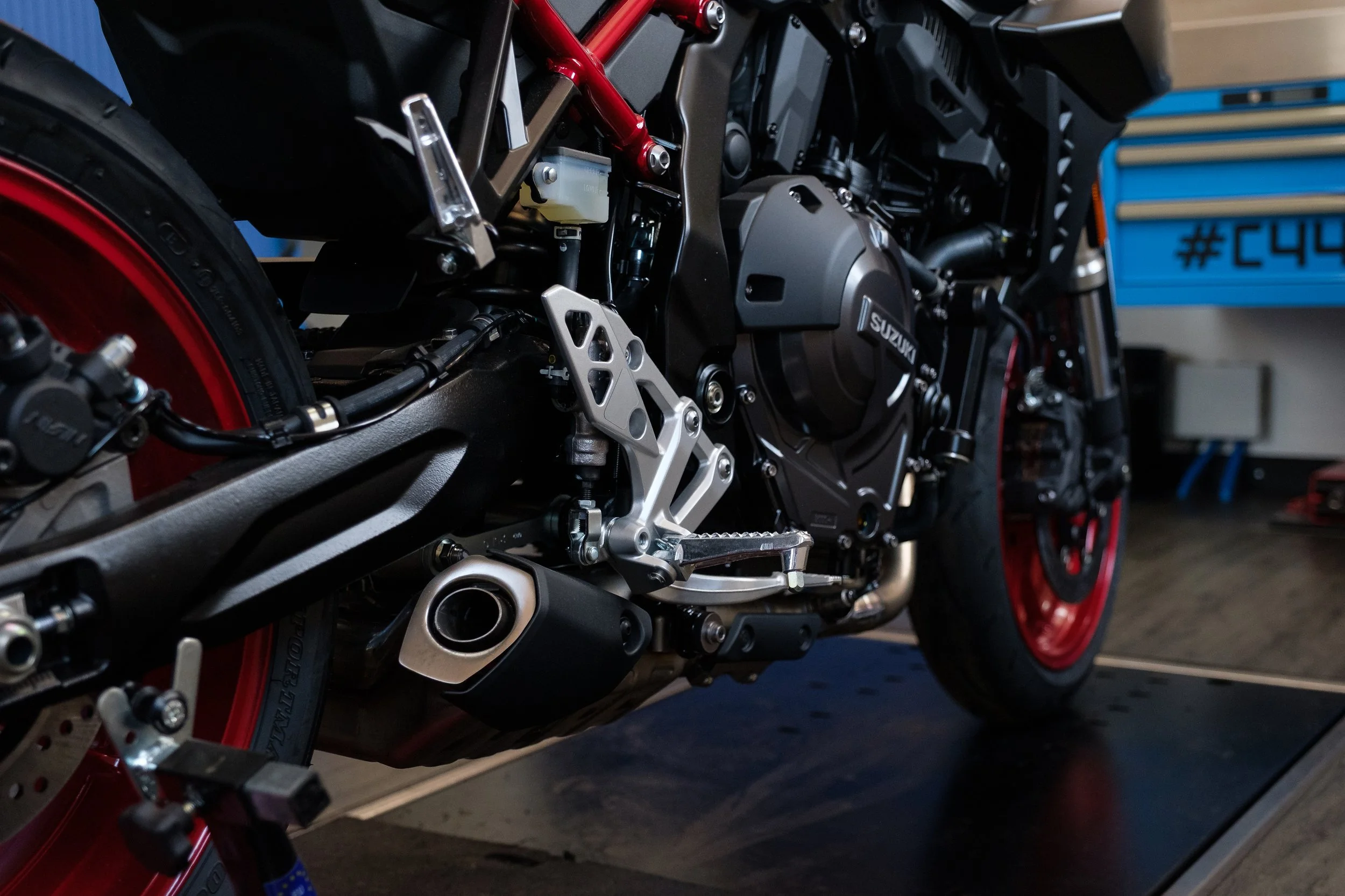 Nahaufnahme eines schwarzen Motorrads mit rotem Rahmen, Fokus auf den Antrieb und das Pedal, im Hintergrund Werkzeugschränke