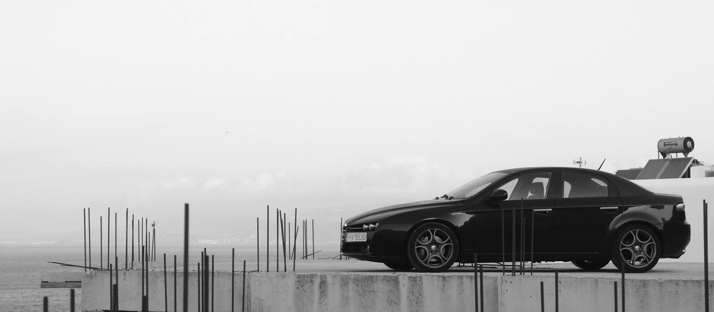 Schwarzer Sportwagen auf einer Brüstung, mit einer Meeres- oder Seenlandschaft im Hintergrund, überwiegend im Schwarz-Weiß-Stil.