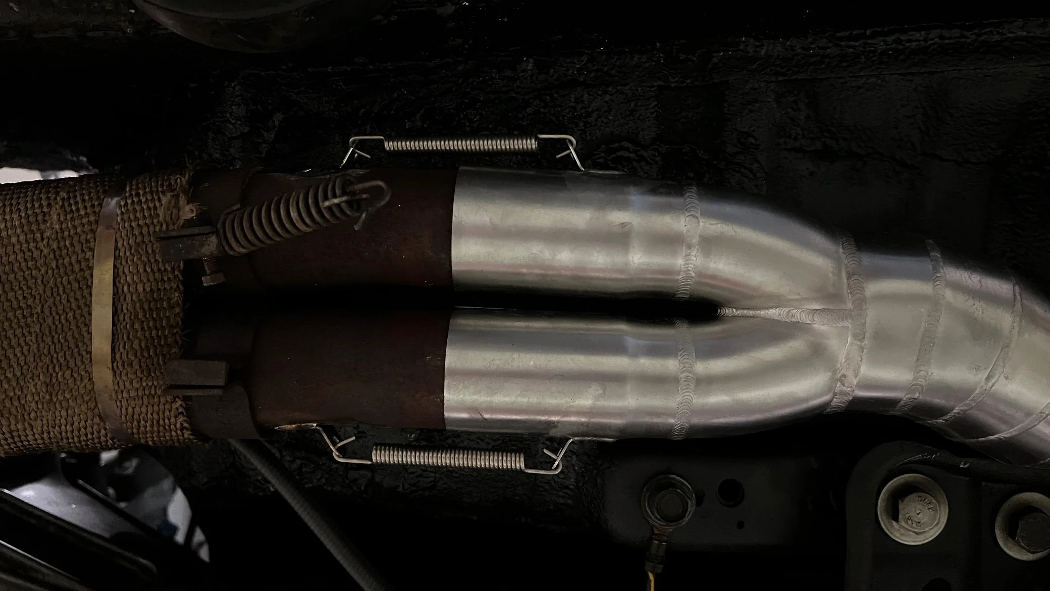 Ein Metallrohr mit mehreren Schweißnähten, umgeben von schwarzen Oberflächen, mit Federn und einem Schutzgitter an der Seite.