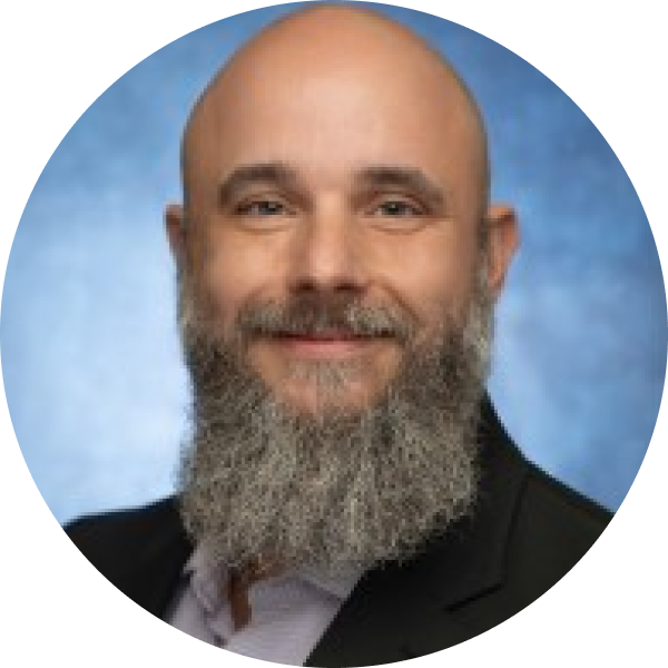 Portrait of a bald man with a full gray beard, wearing a suit, smiling against a blue background.