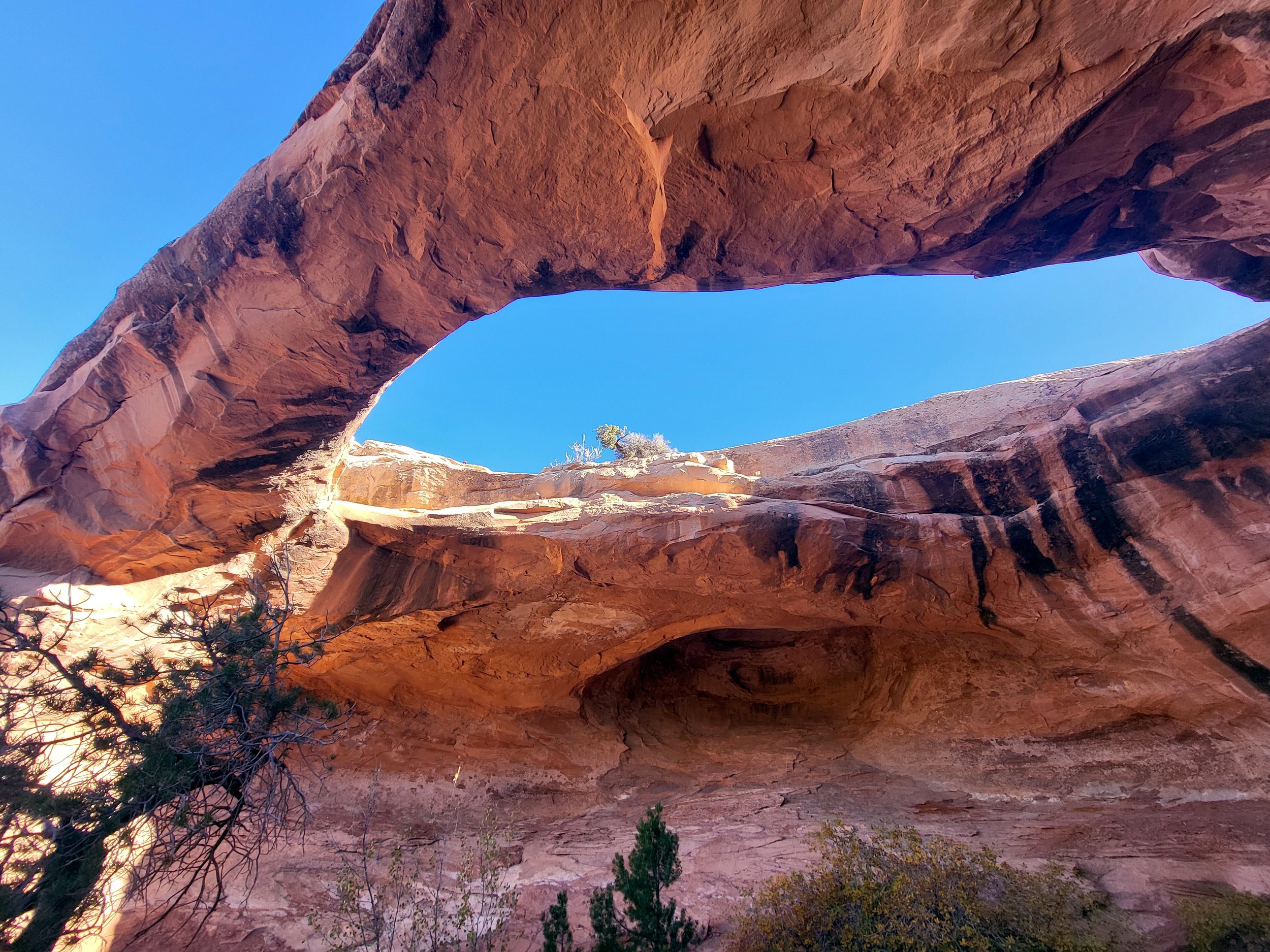 View of a large natural rock arch formation in a desert landscape with clear blue sky and sparse vegetation.