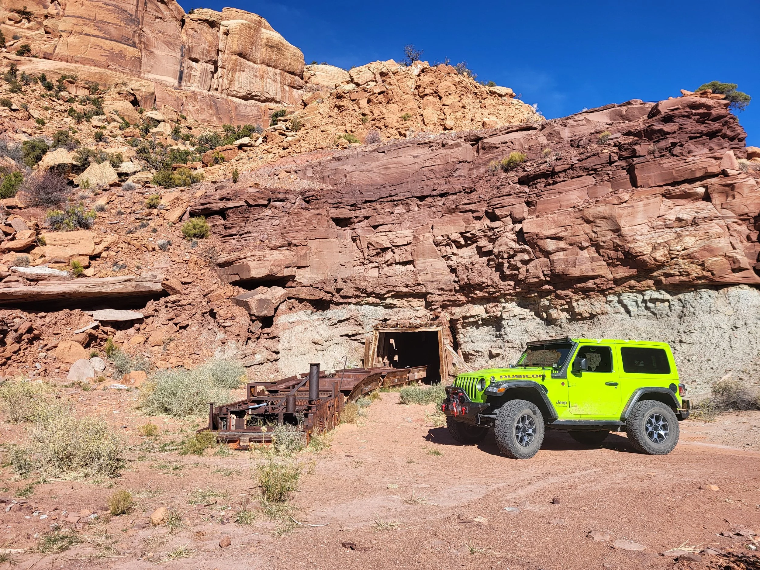 A bright yellow Jeep parked in front of a rocky cliff face with a dark entrance to a mine or tunnel. The landscape is desert-like with low vegetation and a clear blue sky.