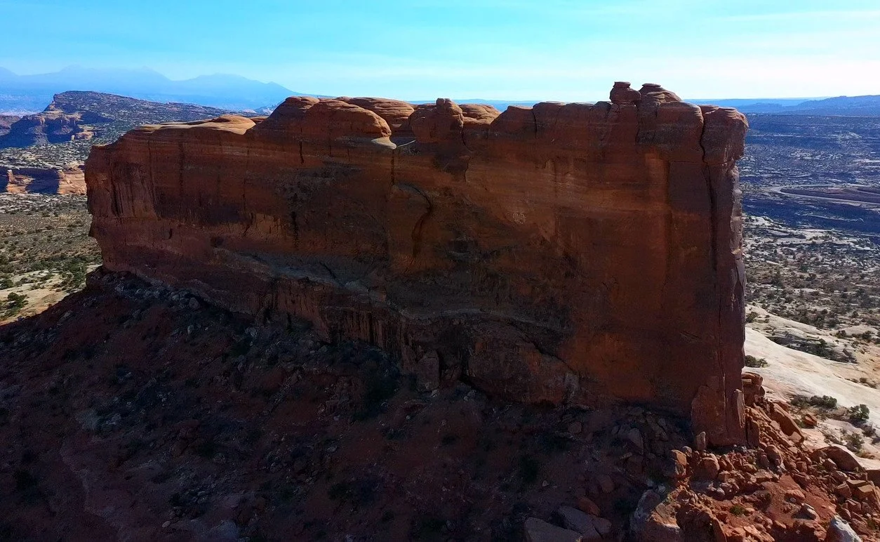 A large red sandstone rock formation on a desert landscape with small town and mountains in the background