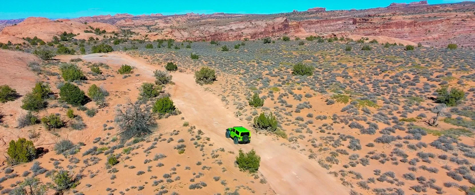 A green vehicle driving on a dirt road through a desert landscape with sparse vegetation and red rock formations in the background.