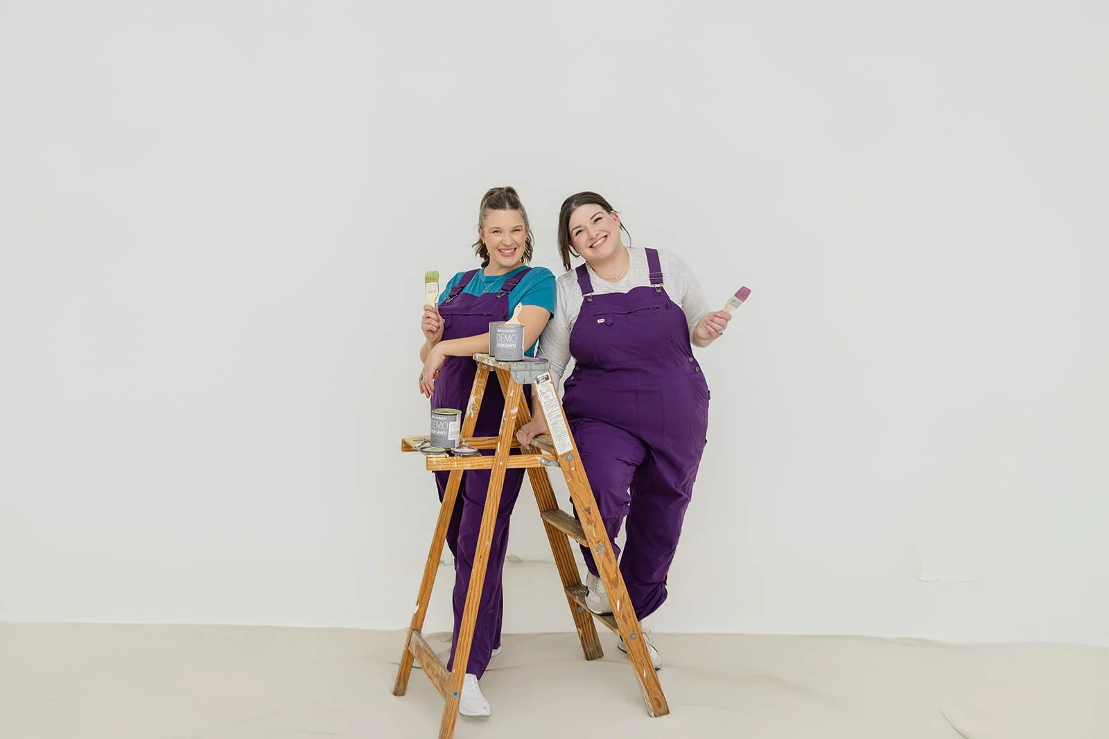 Allie and Leah, owners of The List, standing by ladder with paint brushes in their hands -offering professional wallpaper installation and residential painting services to help MN homeowners with their to-do lists.