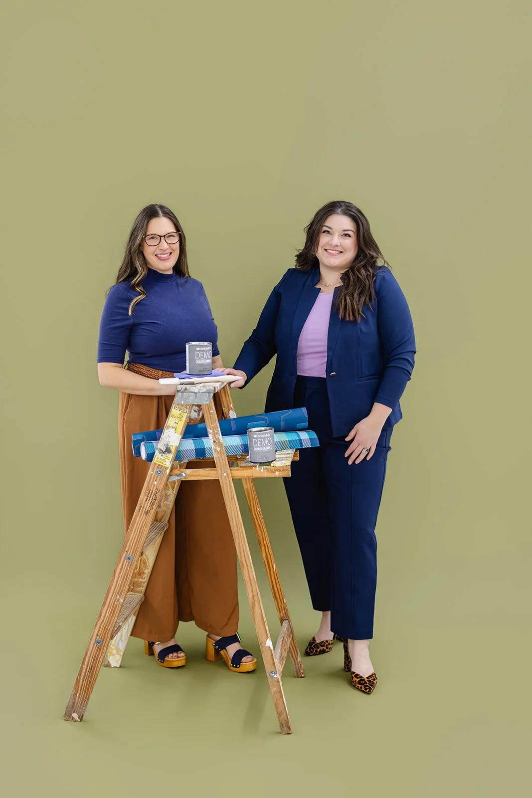 Two women standing by ladder with paint and wallpaper on green background