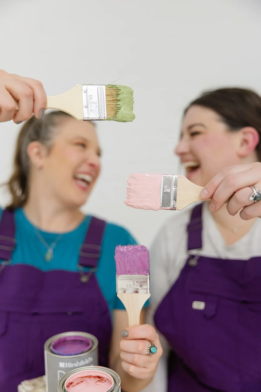 Two women smiling and holding paintbrushes with pink, green, and purple paint, surrounded by open cans of paint.