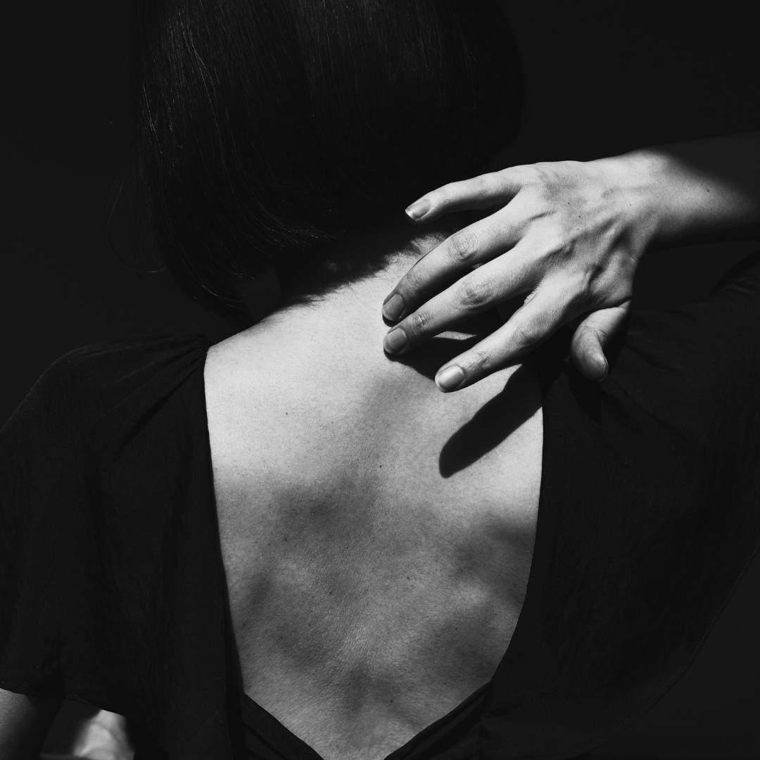 A black and white photograph of a person shown from behind, touching their neck with their right hand, with short dark hair and wearing a dark top.