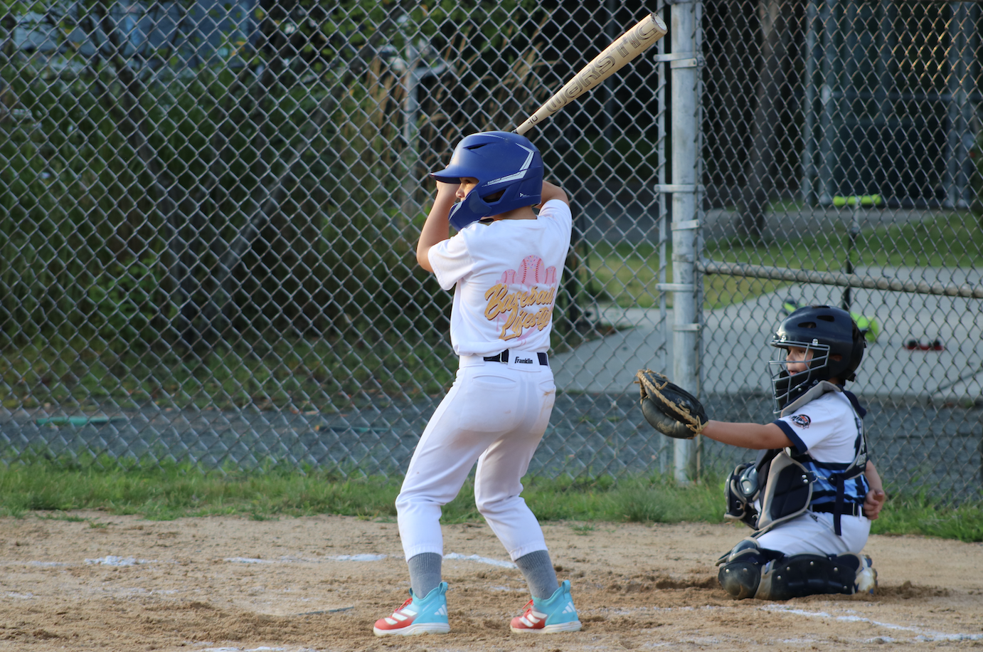 Young baseball player at bat wearing a blue helmet, white uniform, and red and blue shoes, with a catcher in mask and gear crouching behind on a baseball field.
