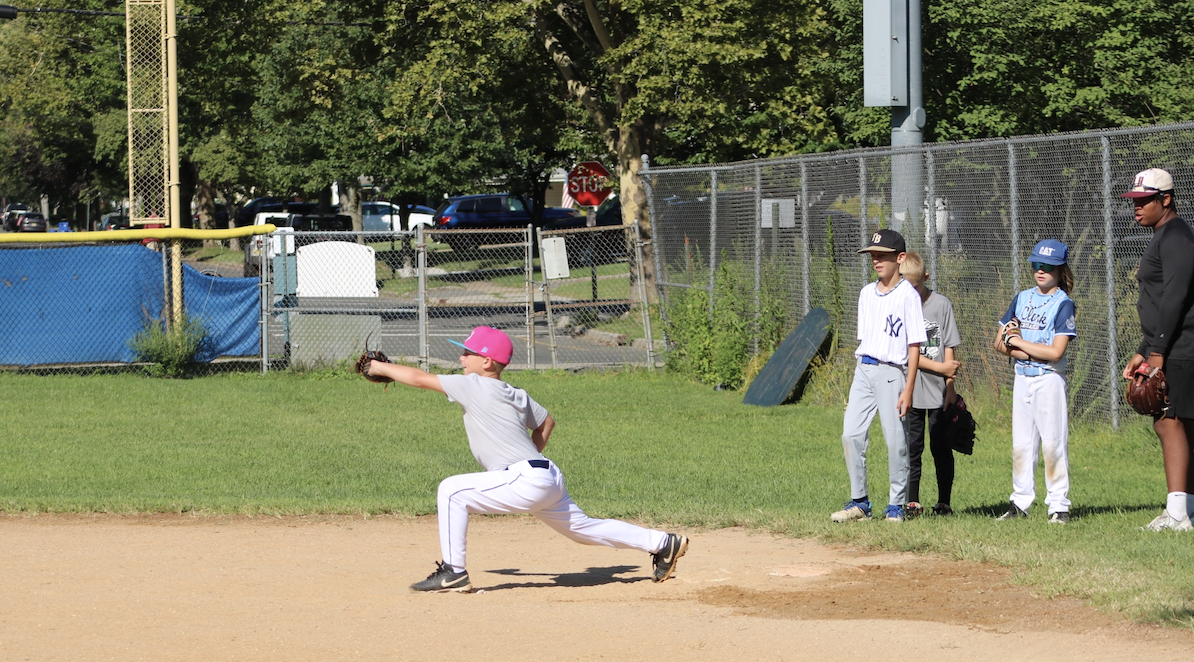 A young baseball player in a gray uniform and pink cap is in a throwing stance on a baseball field, with a group of children and an adult coach watching nearby.