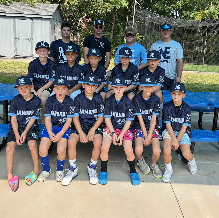 Group of young boys in baseball uniforms and caps posing for a team photo outdoors, with a few adult coaches standing behind them. They are sitting on blue benches at a baseball field with trees in the background.