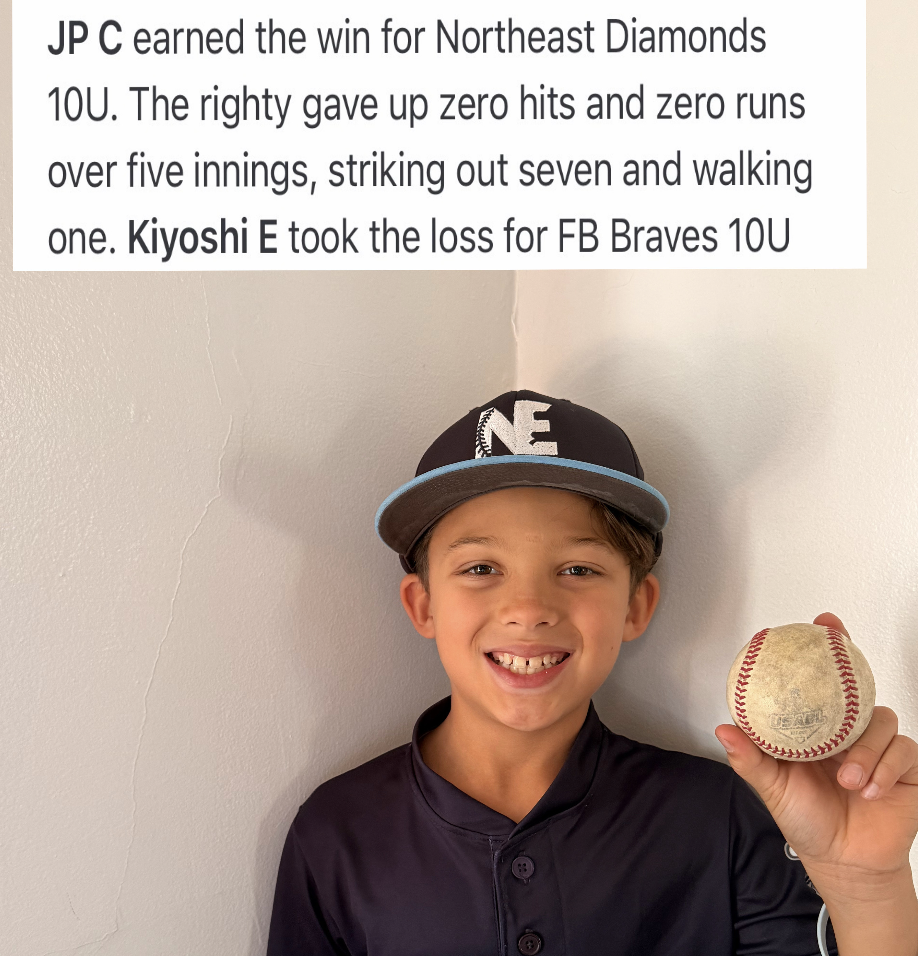 Young boy smiling and holding a baseball, wearing a baseball cap and sports jersey, standing against a plain wall.