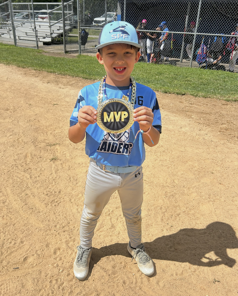 A young boy standing on a baseball field holding an MVP medal, wearing a blue baseball uniform and cap, smiling for the camera.