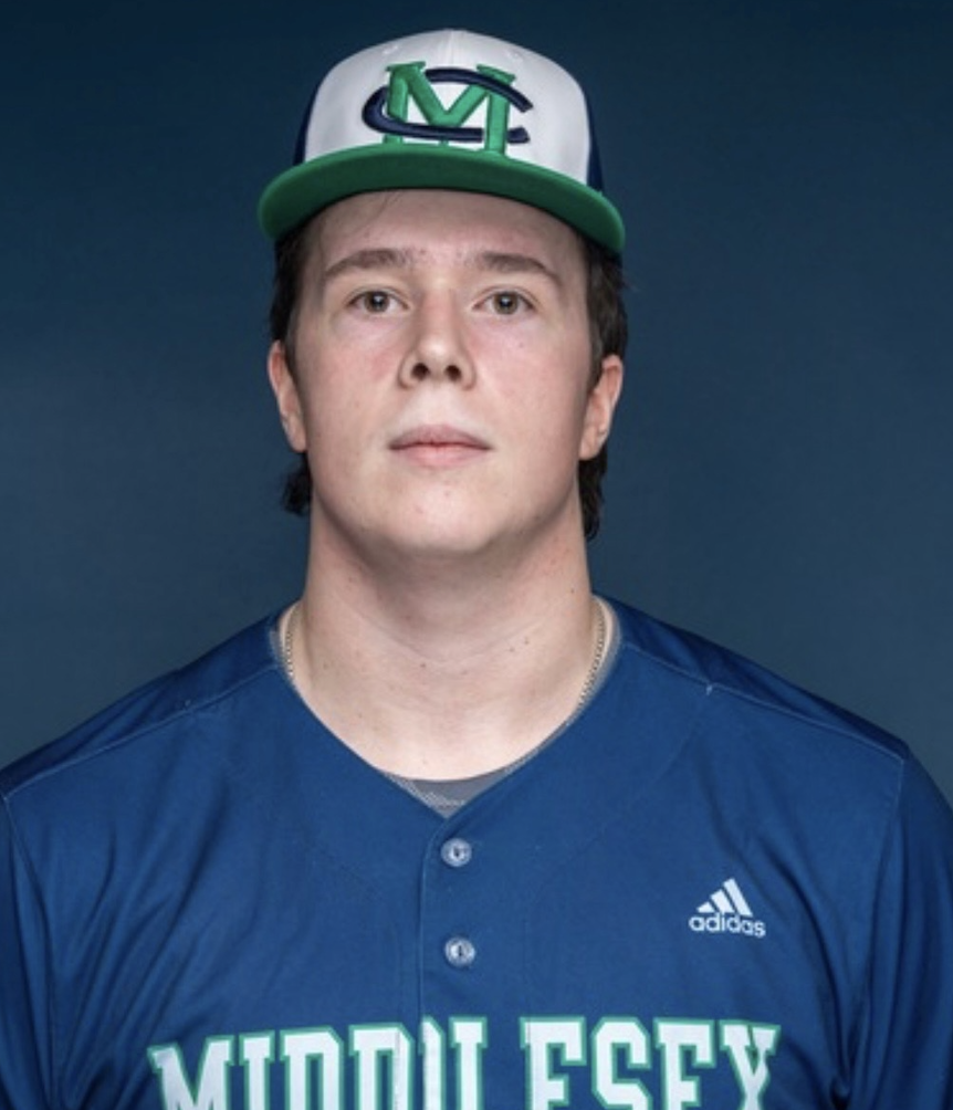 A young man with fair skin and dark brown hair wearing a blue sports jersey with 'MIDDLESSEX' in green and white letters, and a white, green, and black baseball cap with the letter 'M' on it, against a dark background.