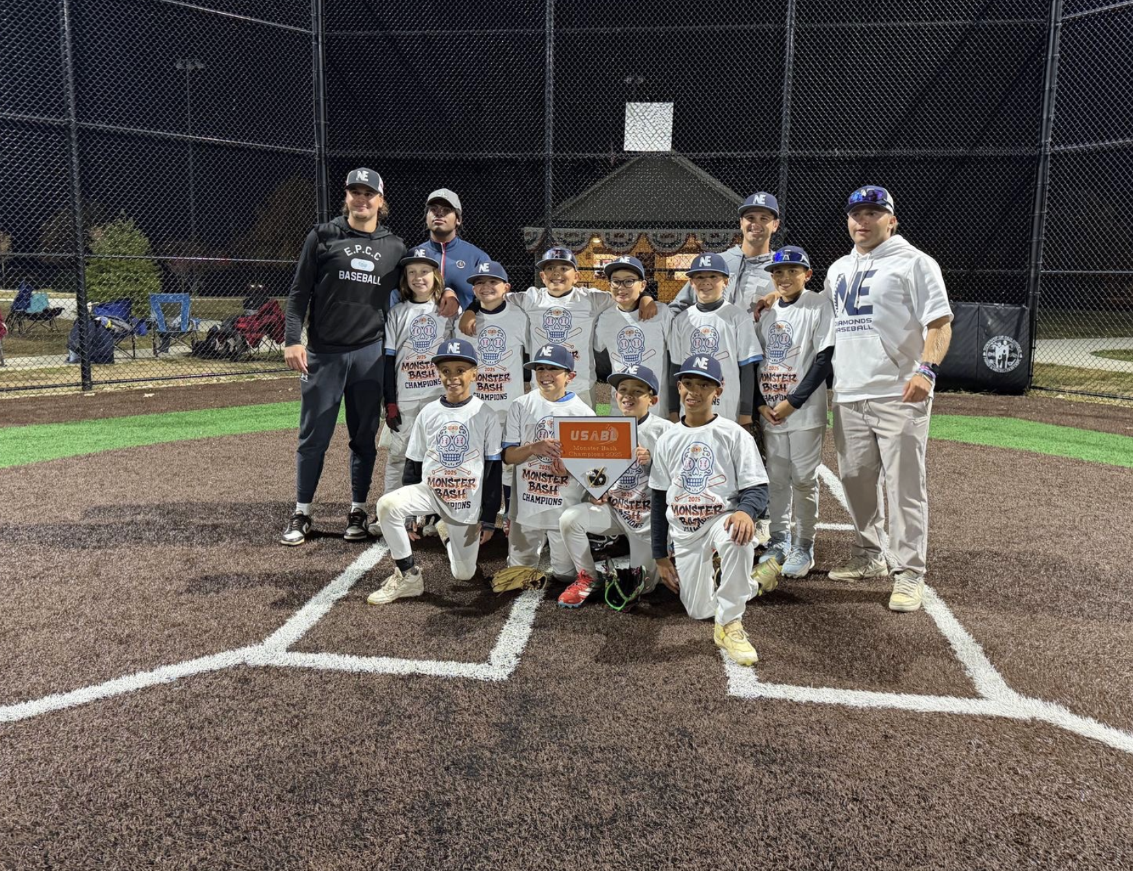 Youth baseball team posing for a photo on a baseball field at night, holding a trophy, with coaches and team members wearing white and blue uniforms, behind a chain-link fence and a carousel in the background.