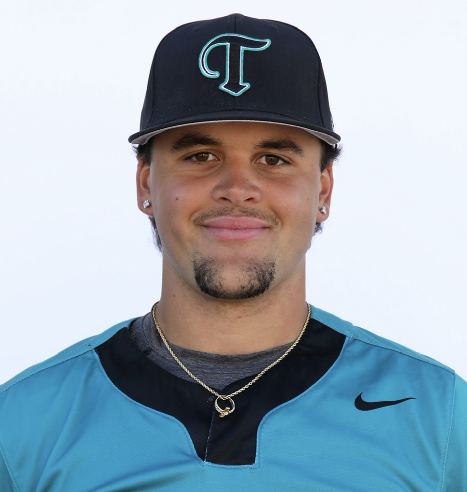 Close-up of a young man wearing a blue sports jersey, a black baseball cap with a turquoise letter T, and silver jewelry against a plain light background.