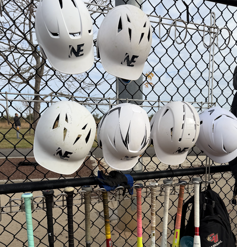 White baseball helmets hanging on a chain-link fence at a baseball field, with baseball bats below and a backpack nearby.