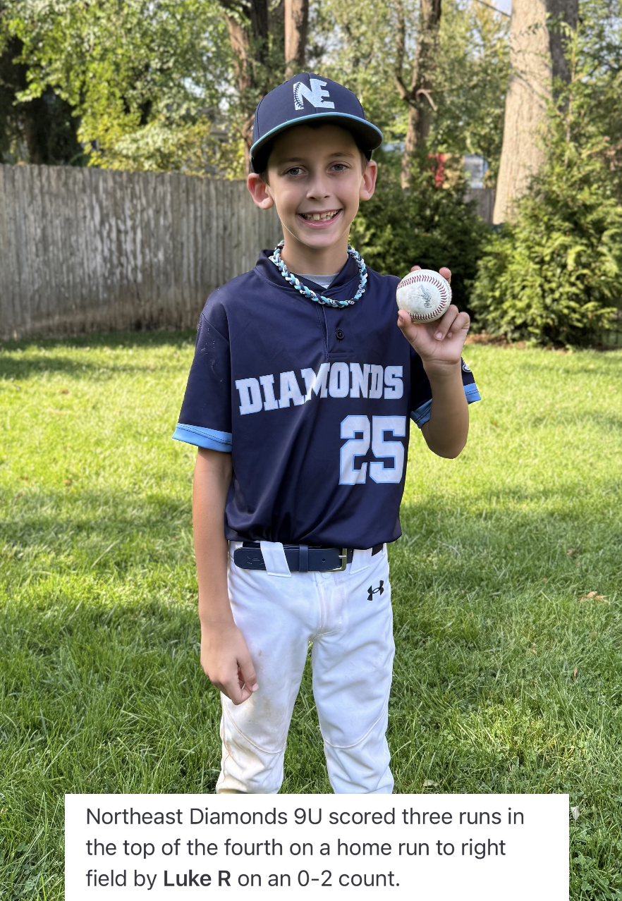 A young boy in a baseball uniform holding a baseball in a backyard with green grass and trees, smiling at the camera.