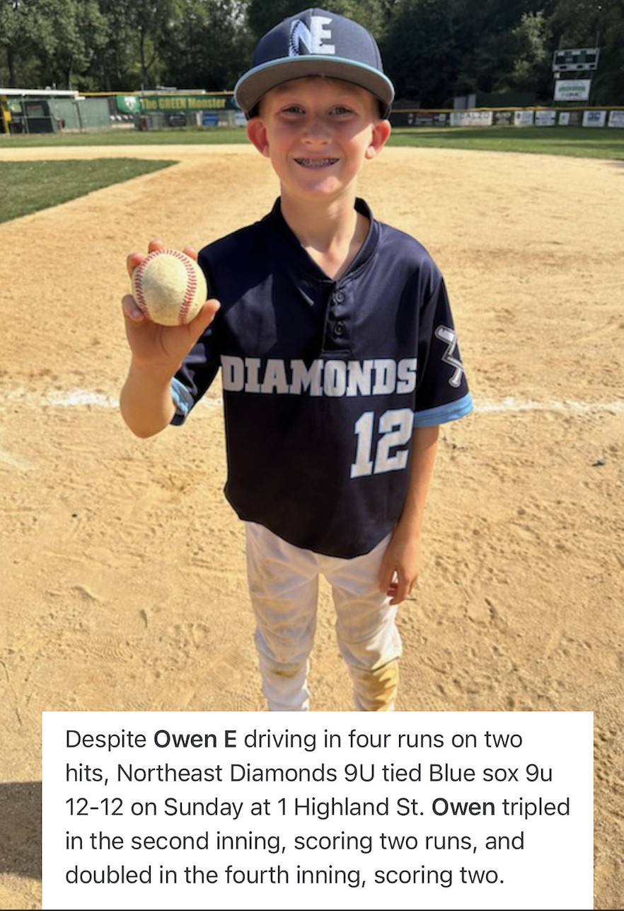 Young boy standing on a baseball field holding a baseball in his right hand, wearing a black jersey with 'DIAMONDS 12' written on it, a gray cap, and beige pants. He is smiling with braces. The background shows the baseball field and trees.