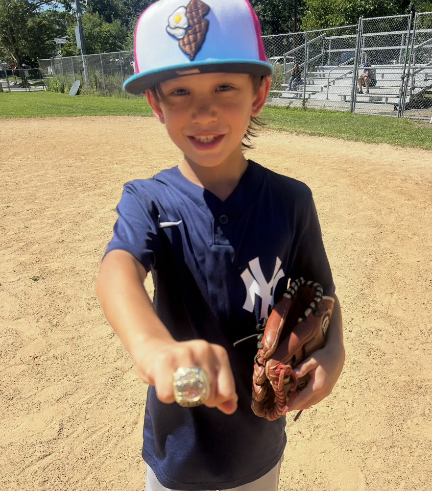 A young boy wearing a New York Yankees baseball jersey and a colorful cap with ice cream and waffle cone embroidered on it, standing on a baseball field. He is smiling and pointing towards the camera, holding a baseball glove and wearing a large ring