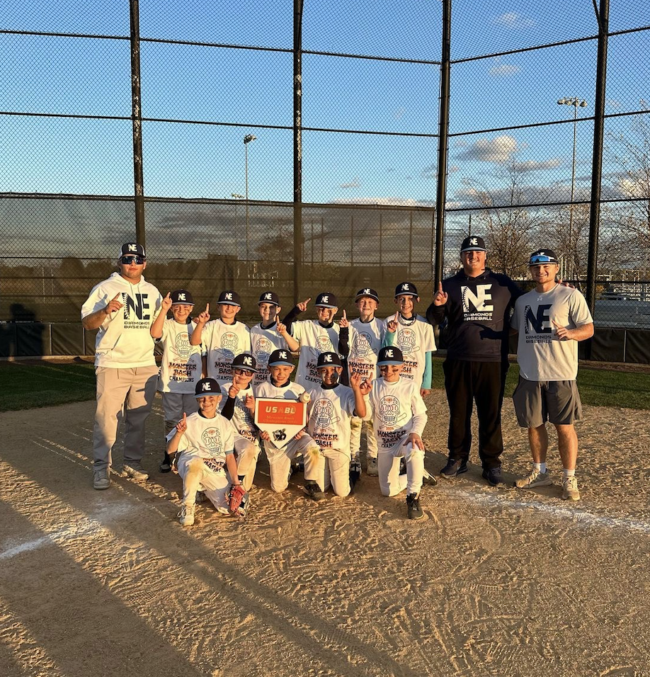 Youth baseball team and coaches celebrating on the field after a game, holding a plaque, all wearing team apparel and caps, with baseball diamond and chain-link fence in the background.