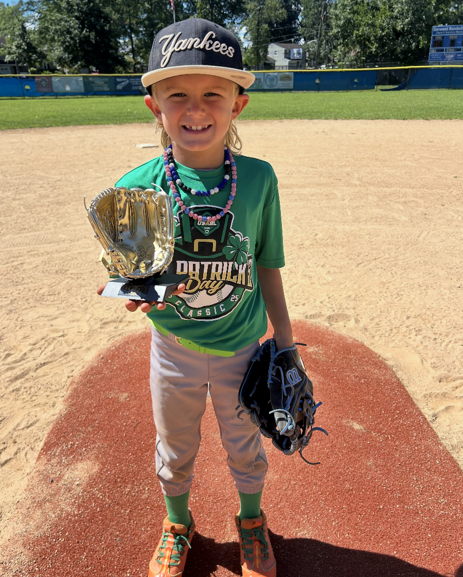 A young boy wearing a Yankees cap, green baseball jersey, khaki pants, and orange cleats standing on the pitcher's mound of a baseball field. He is smiling and holding a gold trophy glove in his right hand and a black baseball glove in his left hand.