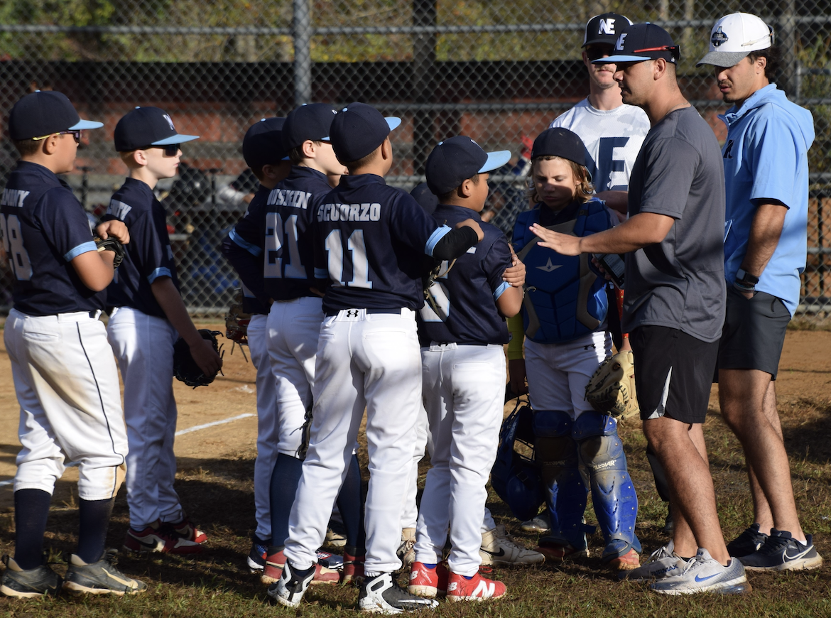 Kids on a baseball team gather around their coach during a game, some wearing uniforms and caps, with a chain-link fence in the background.