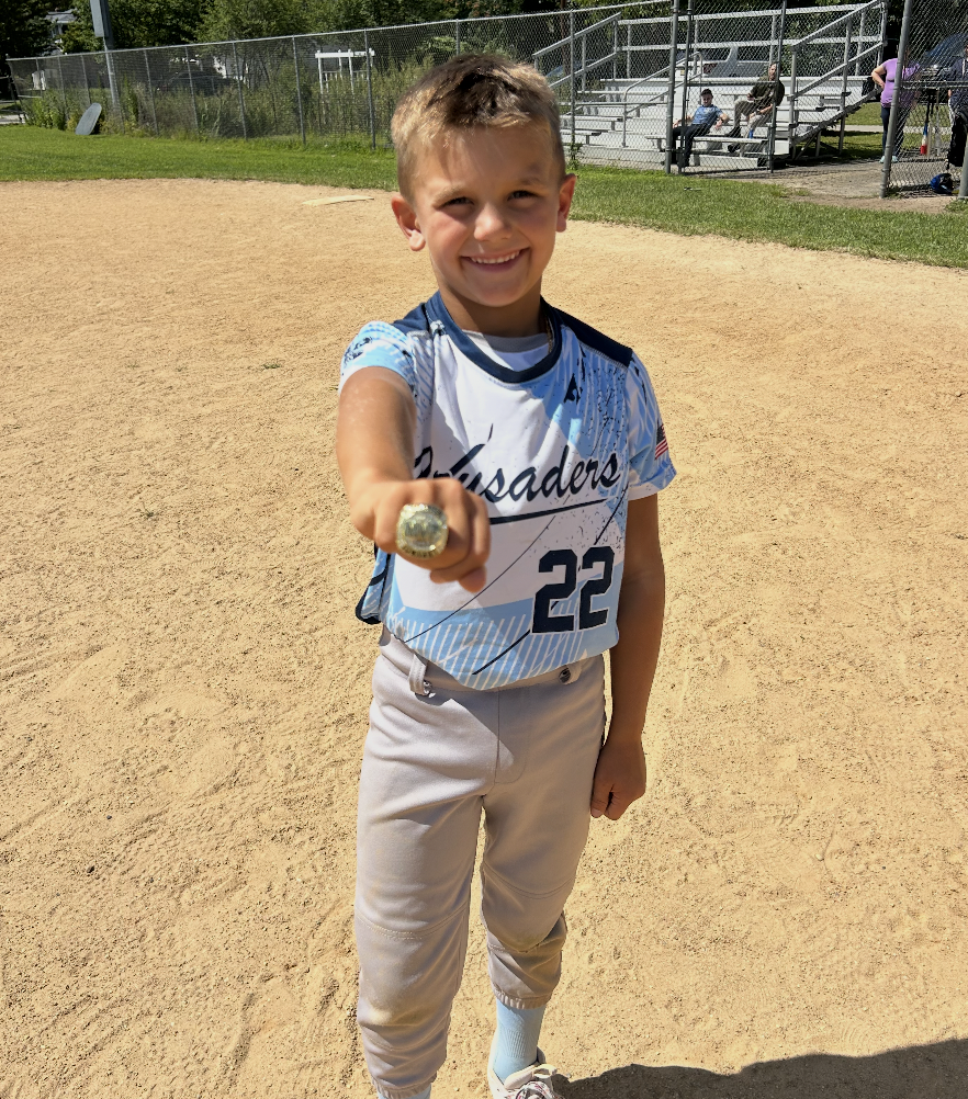 Young boy smiling and showing off a large ring on his finger, standing on a baseball field in a baseball uniform with the number 22 and team name "Crusaders".