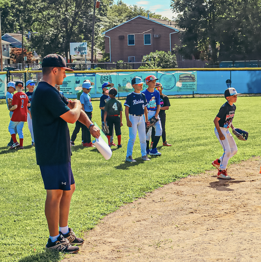 Young baseball players in jerseys and caps on a grassy field with an coach or referee holding a water bottle, other children lined up in the background, and houses and trees in the distance.