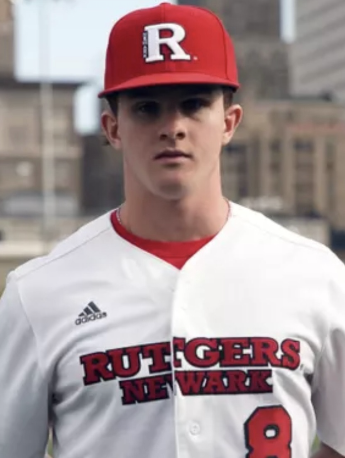 Young man in white baseball uniform with red and black lettering for Rutgers Newark, wearing a red cap with a white 'R' logo.