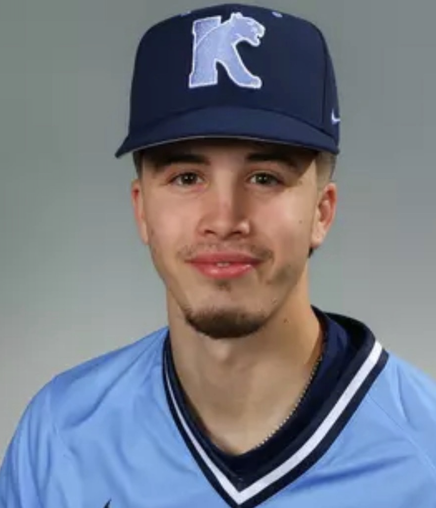 A young man wearing a blue sports cap with a white letter 'K' on it, a blue sports jersey with white and black accents, and a black shirt underneath, smiling at the camera against a plain gray background.