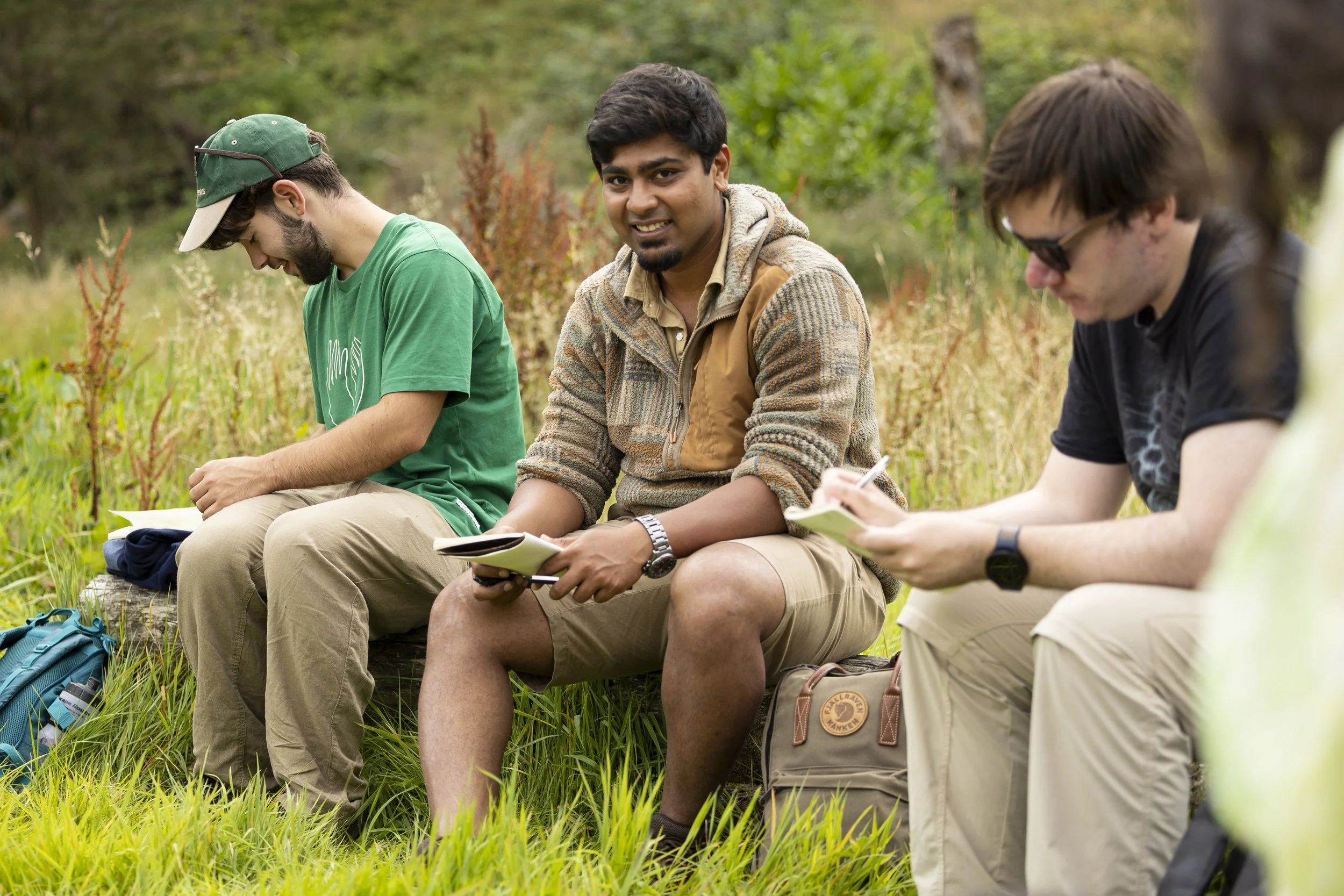 A young man with others around him sat on a log in the countryside with an book in hand for drawing. 