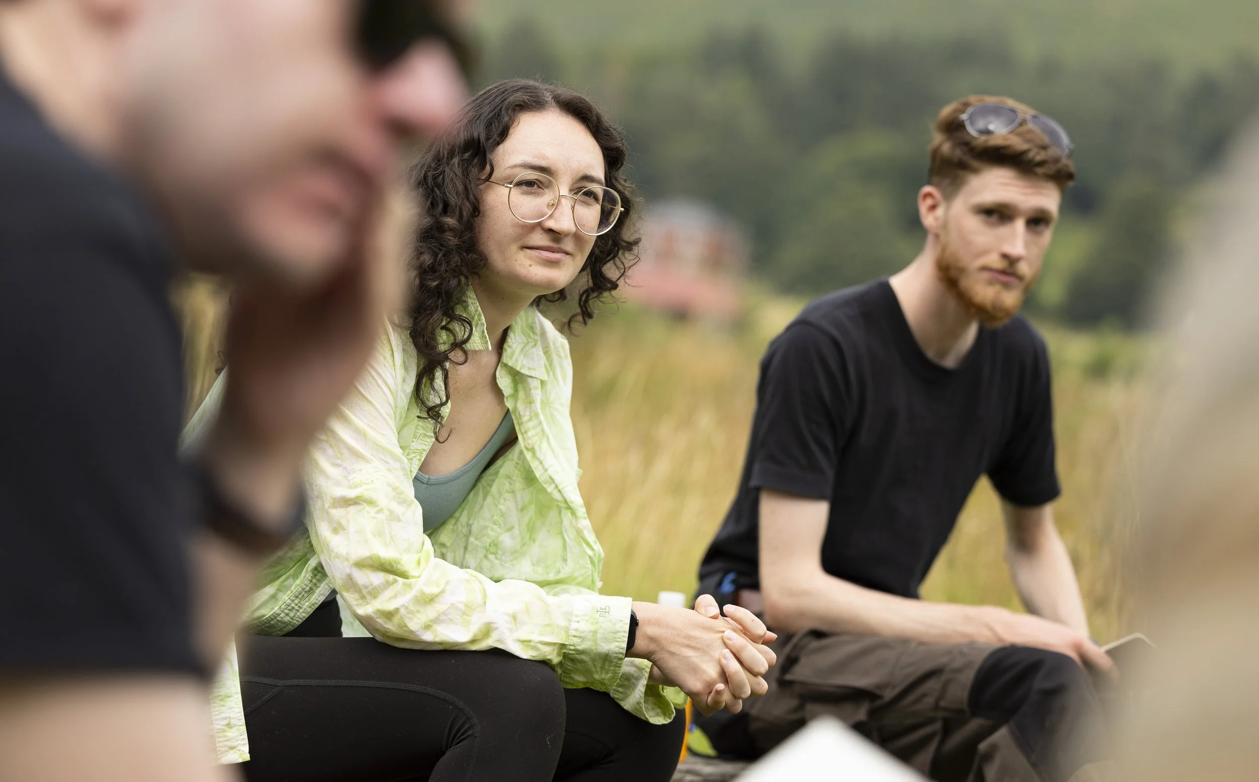 Three people sitting outdoors in a grassy area, engaging in a conversation, with trees and a house in the background.