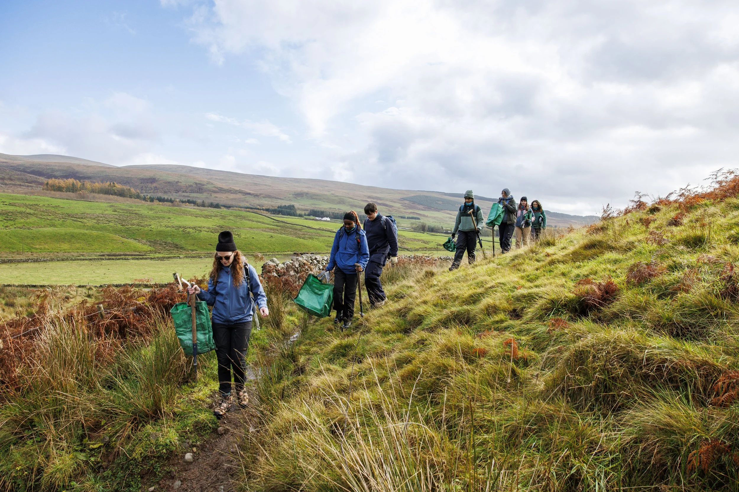A grassy field with a picnic table on the left and a ring of wooden logs on the right. In the background, there are rolling hills with patches of green and brown, trees, a small red building, and a faint rainbow. The sky is partly cloudy.