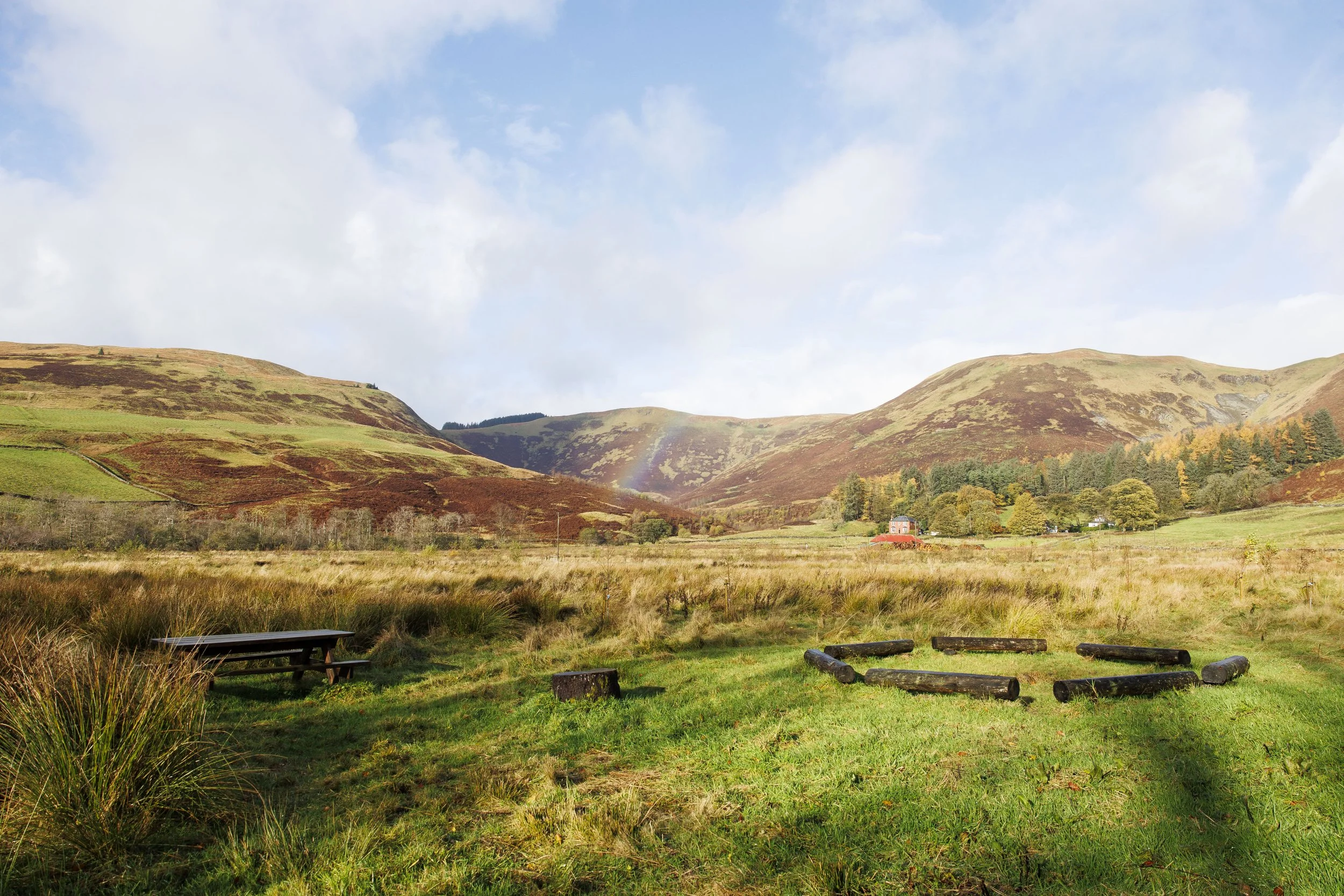 A grassy field with a picnic table on the left and a ring of wooden logs on the right. In the background, there are rolling hills with patches of green and brown, trees, a small red building, and a faint rainbow. The sky is partly cloudy.