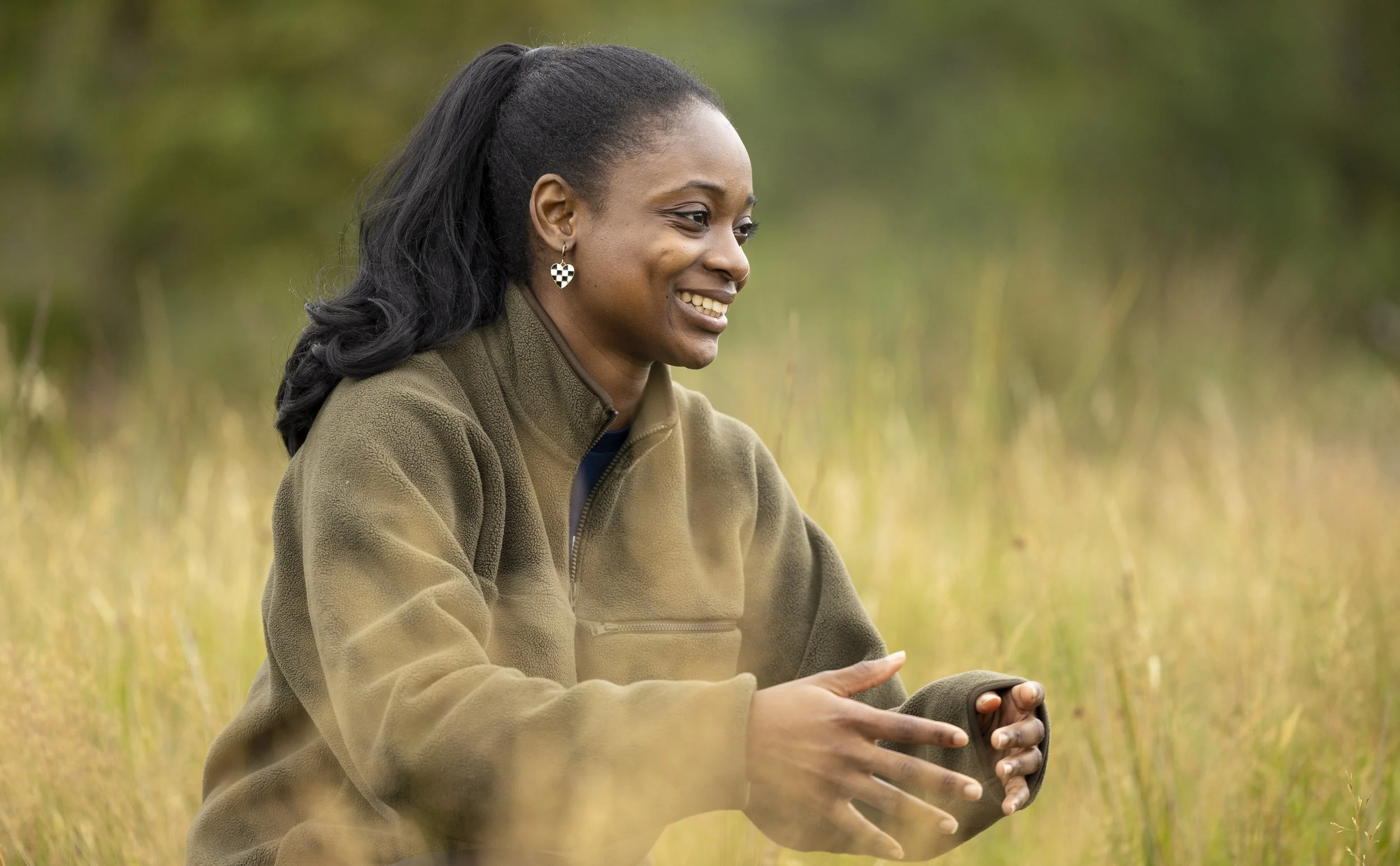 A woman with dark hair in a ponytail, wearing a brown fleece jacket, sitting outdoors in a grassy field, smiling and holding her hands together.